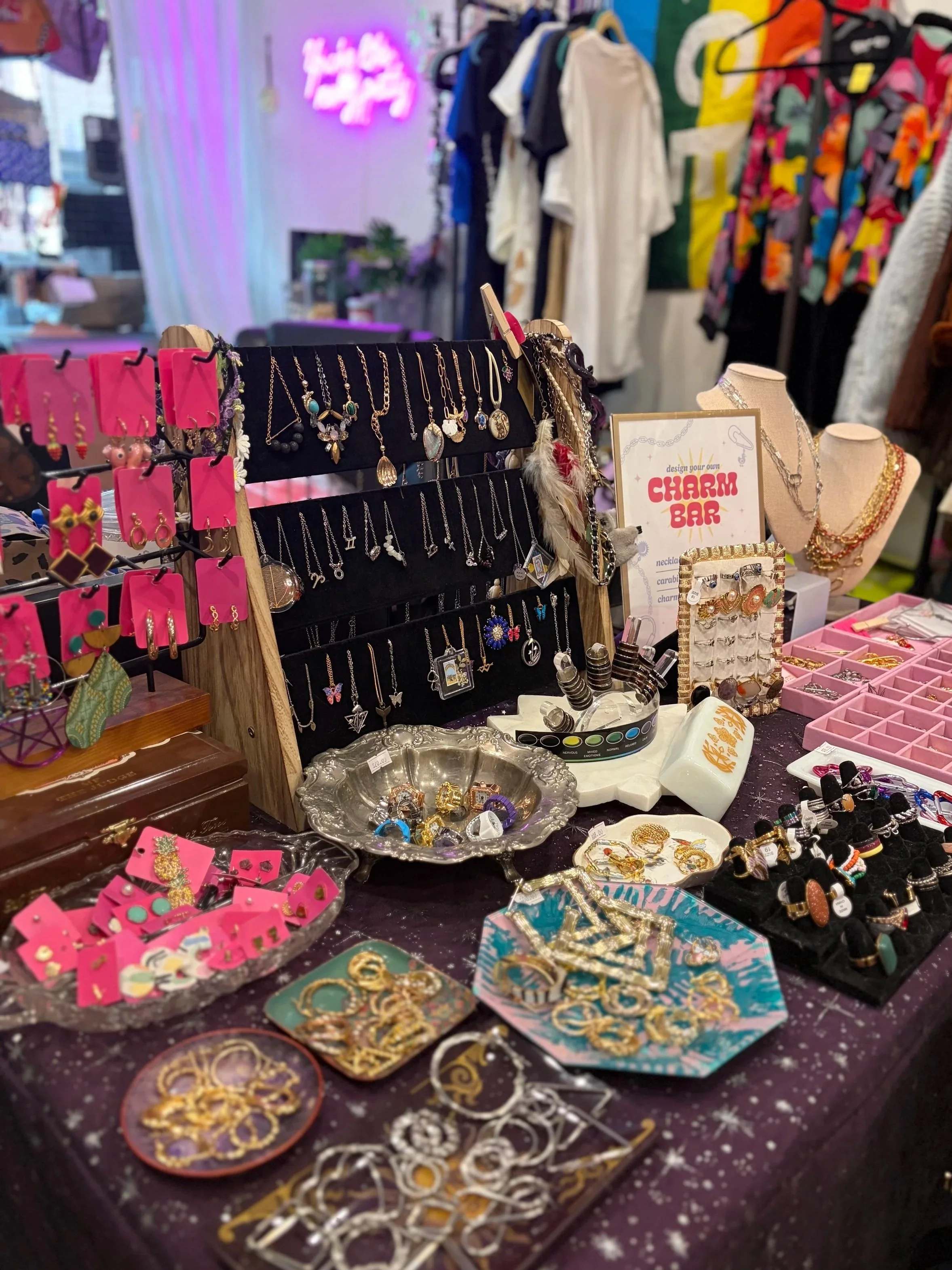 A jewelry and accessories stall with necklaces, earrings, rings, and bracelets displayed on black and pink cards, decorative trays, and stands at an indoor market, with clothing hanging in the background.