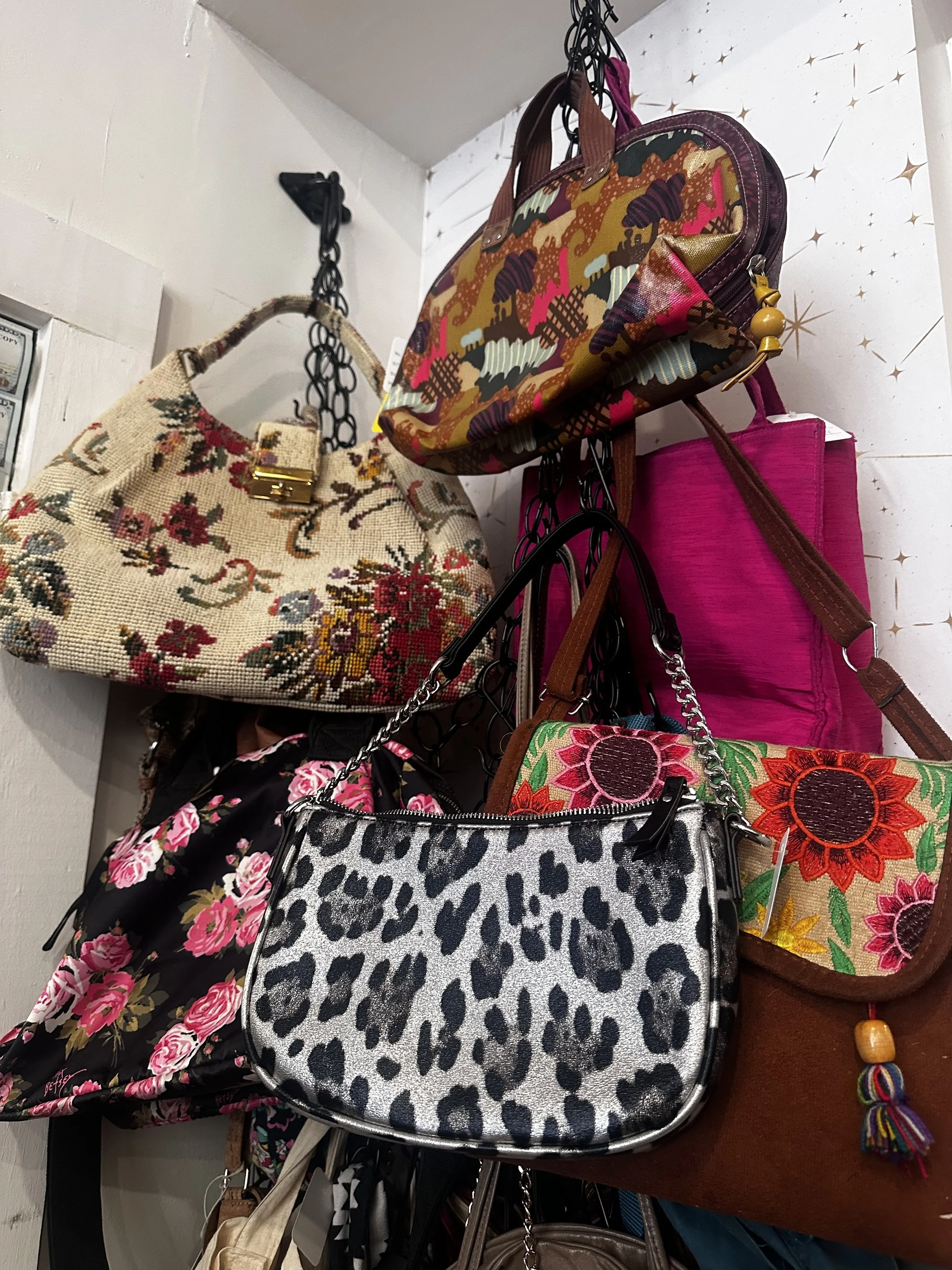 Assorted handbags hanging on hooks against a white wall with star pattern, including a floral tapestry bag, a leopard print bag, a pink bag, and a colorful embroidered bag.