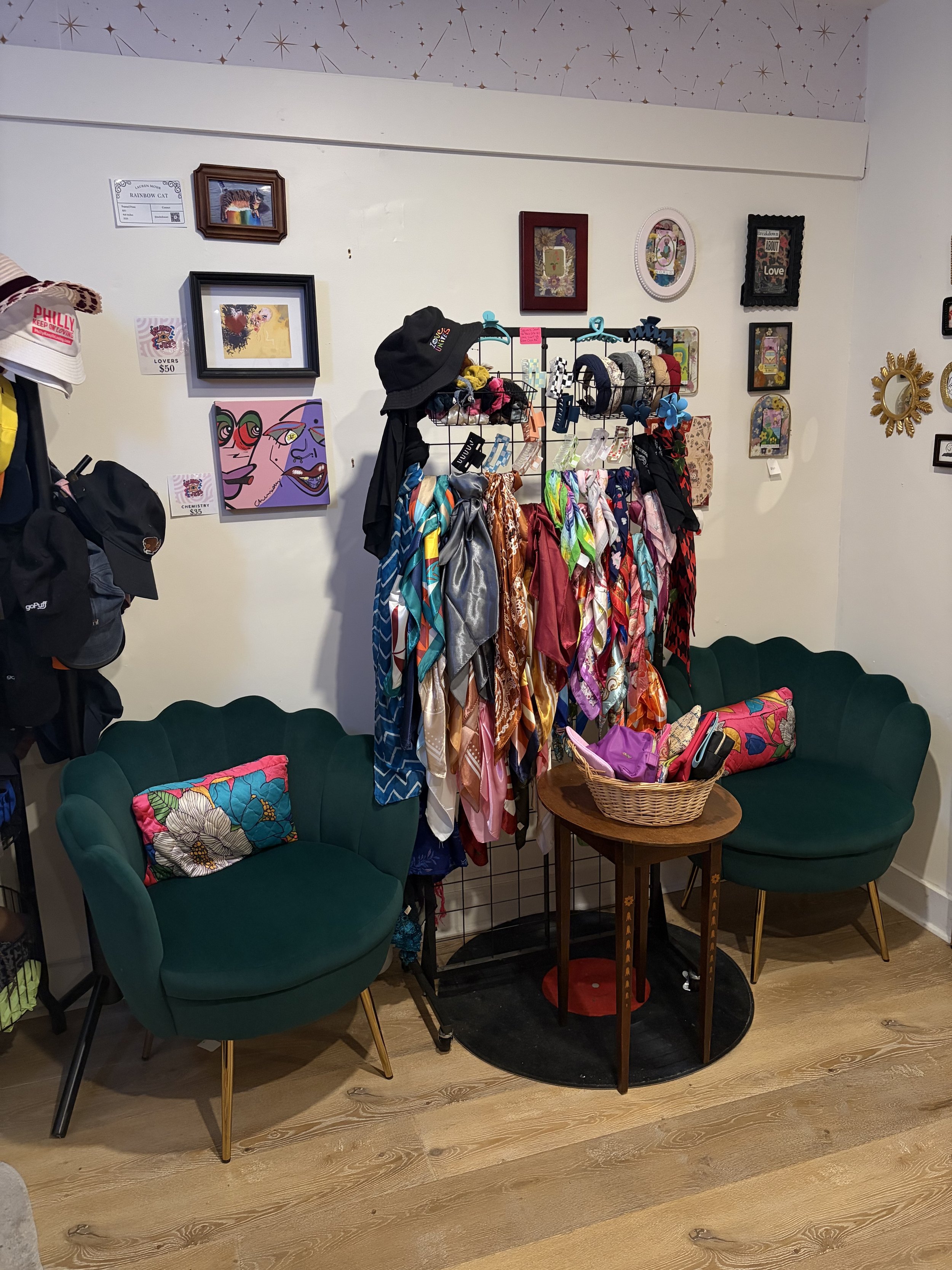 A cozy corner with two green velvet chairs with floral pillows, a small round wooden table with a basket and some items, a clothing rack with scarves and hats, and wall art including framed pictures and artwork.