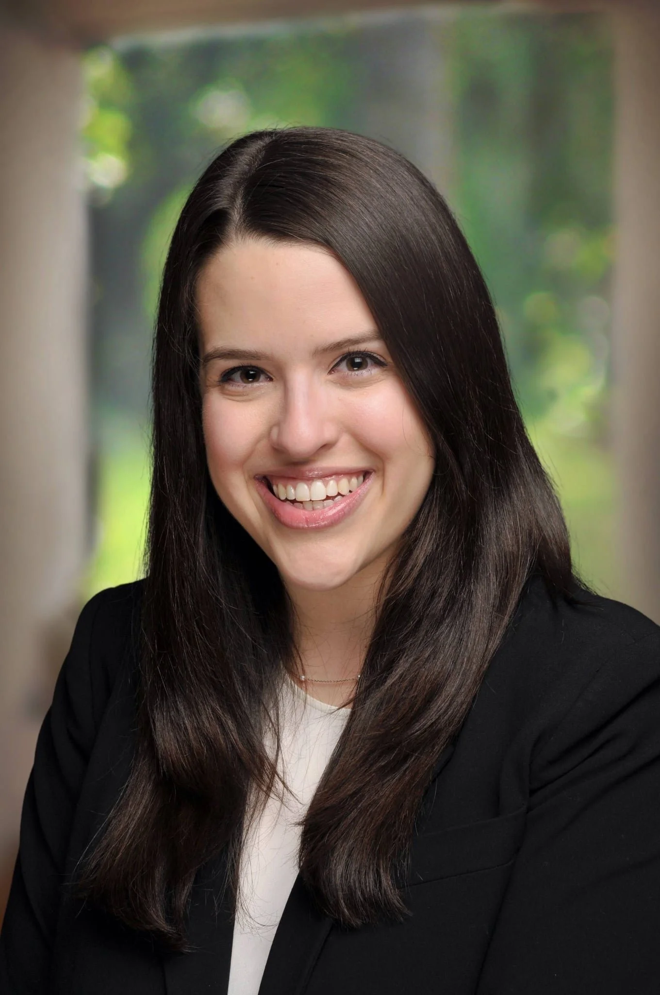 A young woman with long dark brown hair, smiling and looking directly at the camera, wearing a black blazer and white top, with a blurred green outdoor background.