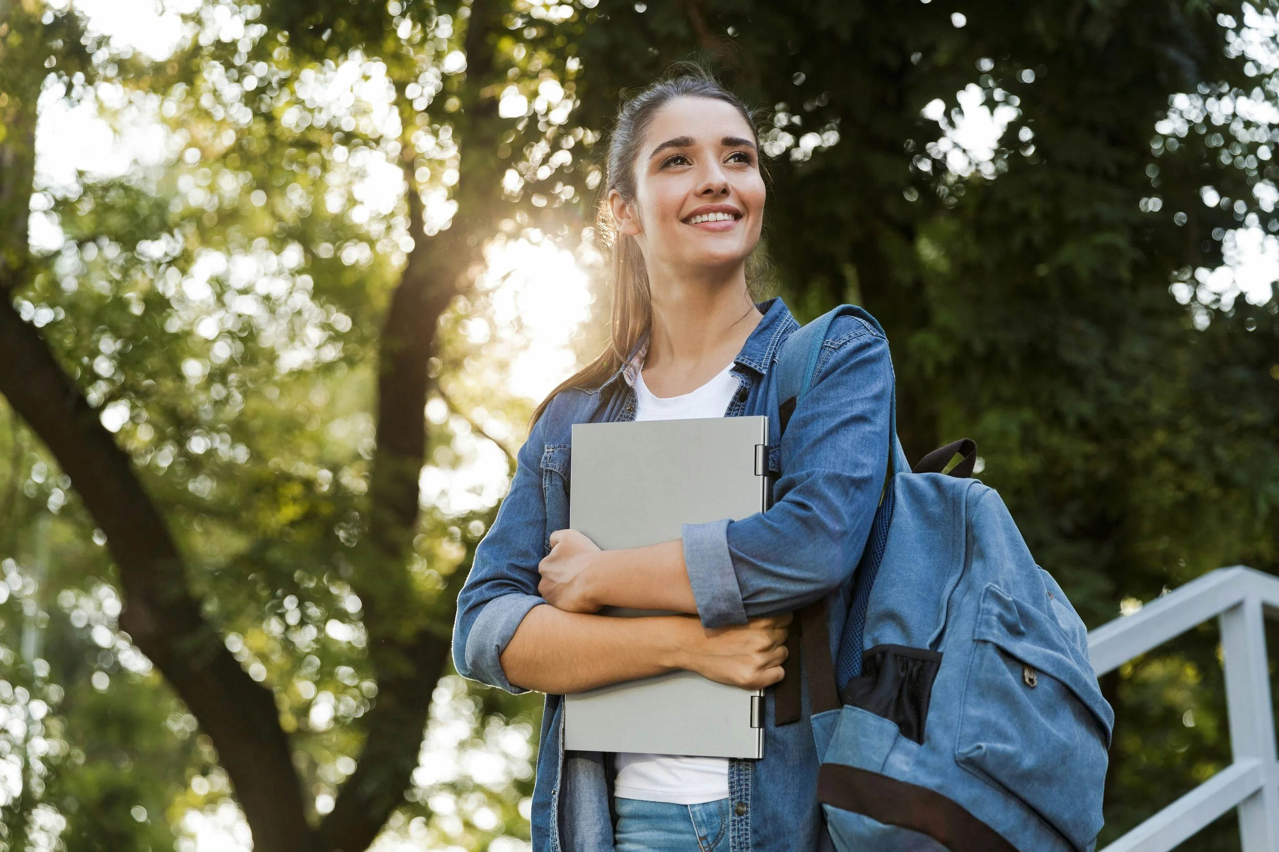 A young woman with long hair, smiling, holding a notebook in front of her chest, standing outdoors under a tree with sunlight filtering through the leaves, wearing a denim jacket and carrying a backpack.