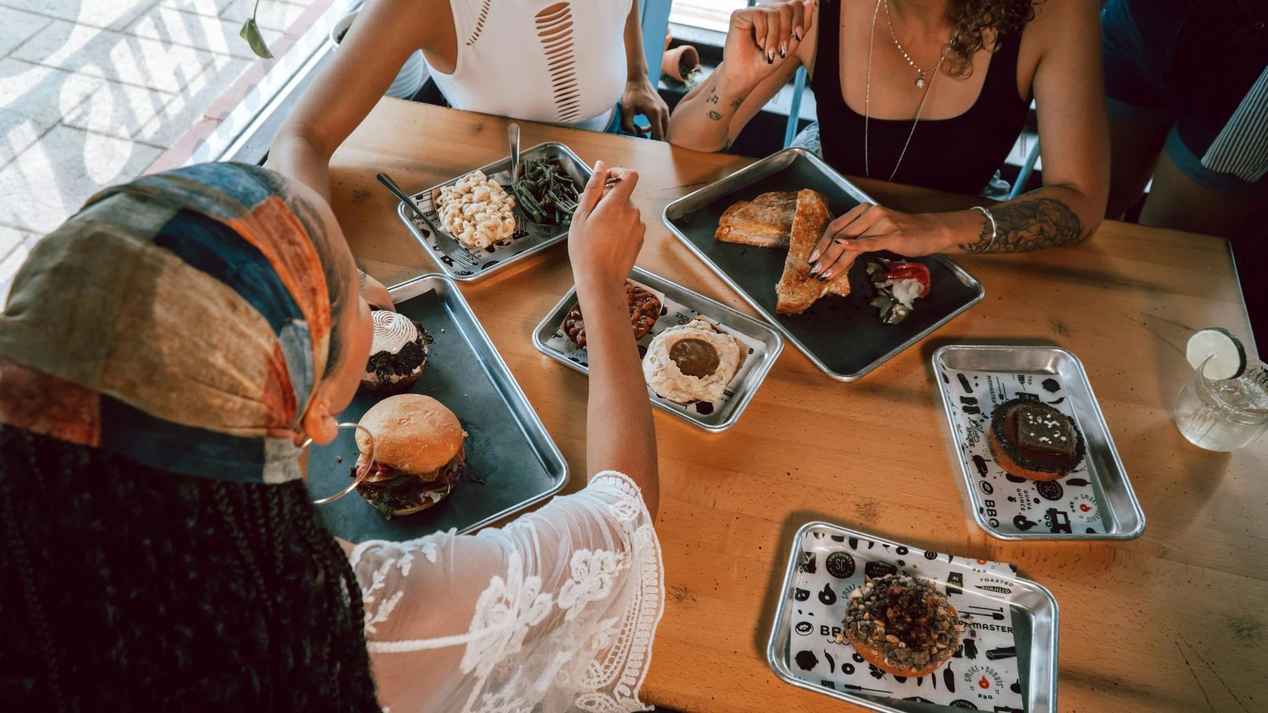 People sitting around a wooden table with metal trays of food, including a burger, fried food, a slice of pizza, and desserts, in a casual restaurant setting.