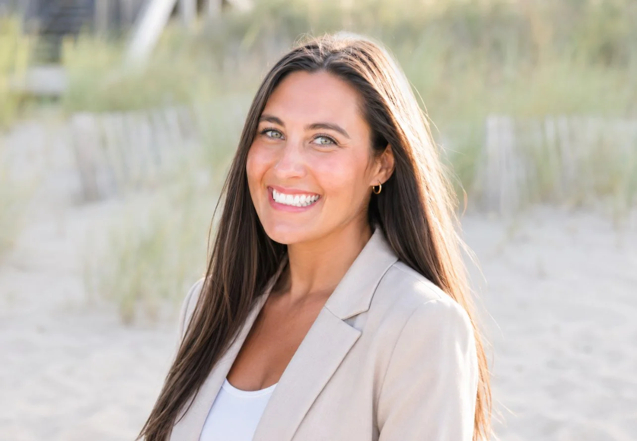 A smiling woman with long brown hair, wearing a beige blazer and white top, standing outdoors at the beach during daytime.