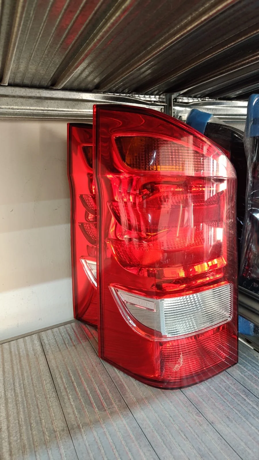 A set of red taillights, likely from a Mercedes Benz, leaning against a wall on a wooden shelf in a store or warehouse.