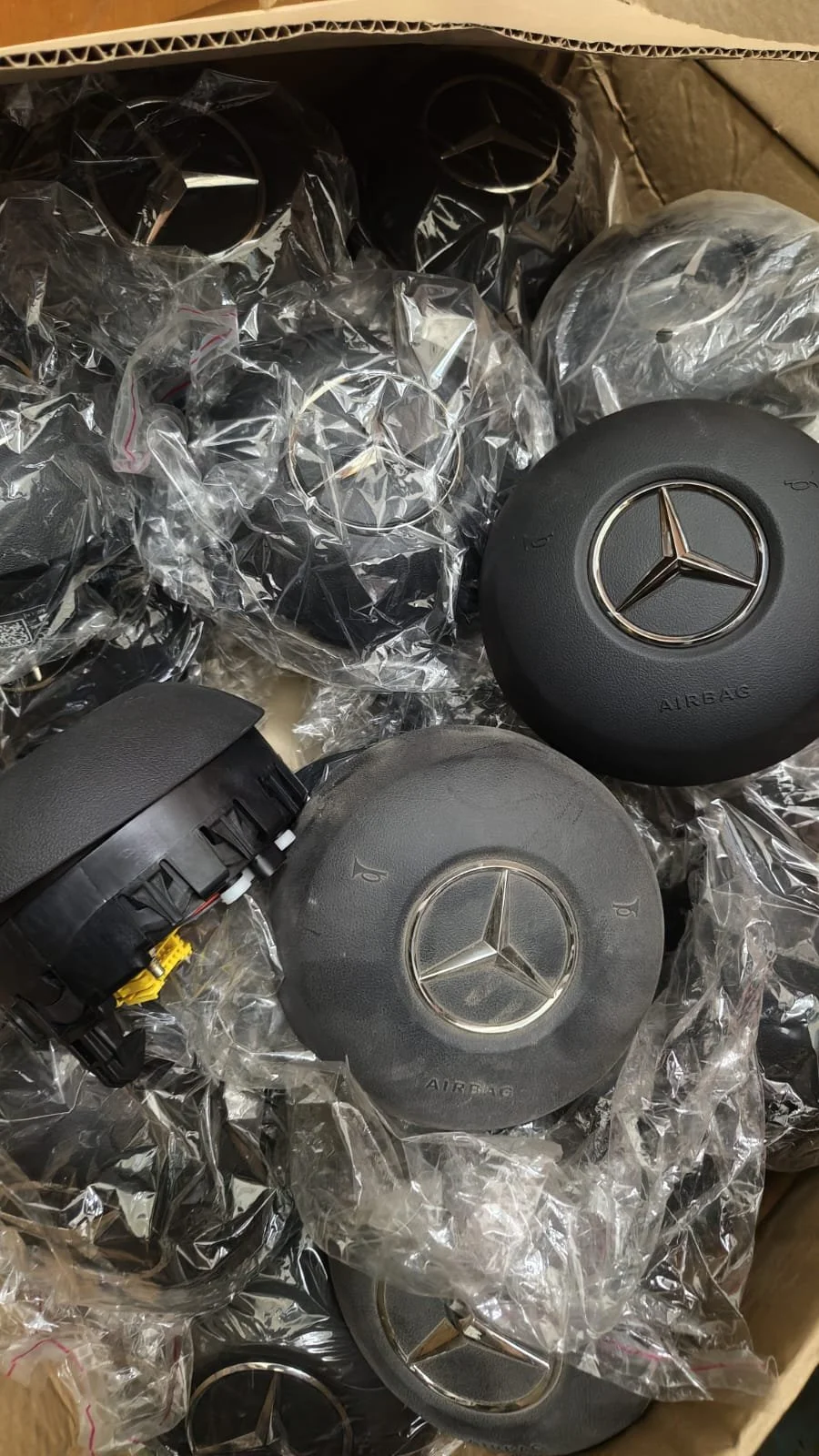 Several Mercedes-Benz steering wheel airbags with the Mercedes logo, some in plastic packaging, in a cardboard box.