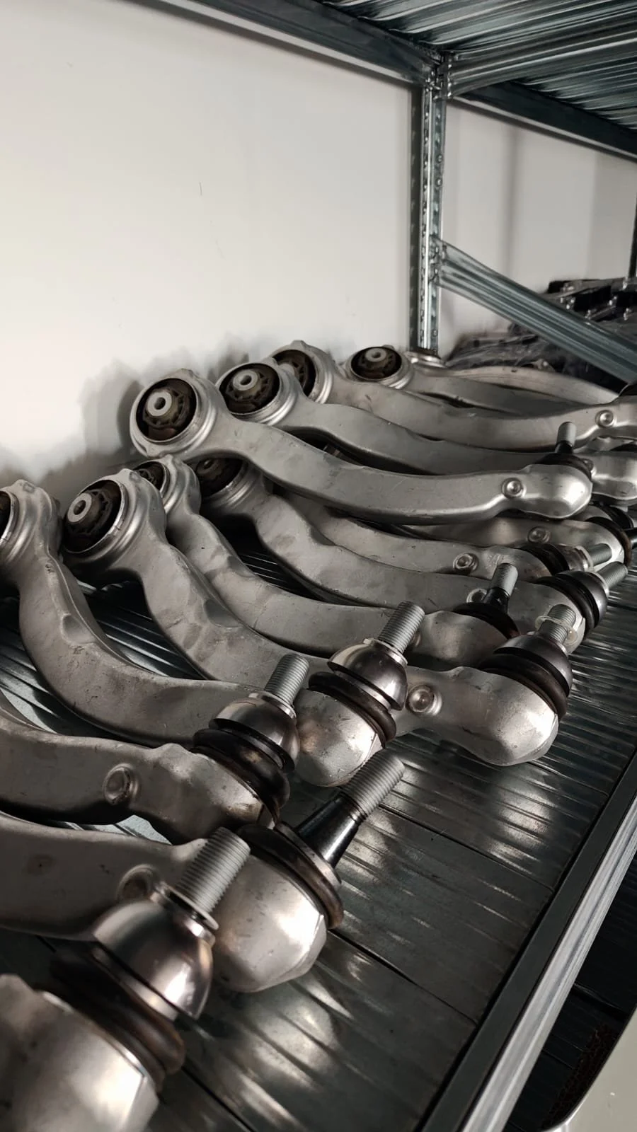Multiple automotive control arms with ball joints stored on a metal shelf in a workshop or storage area.