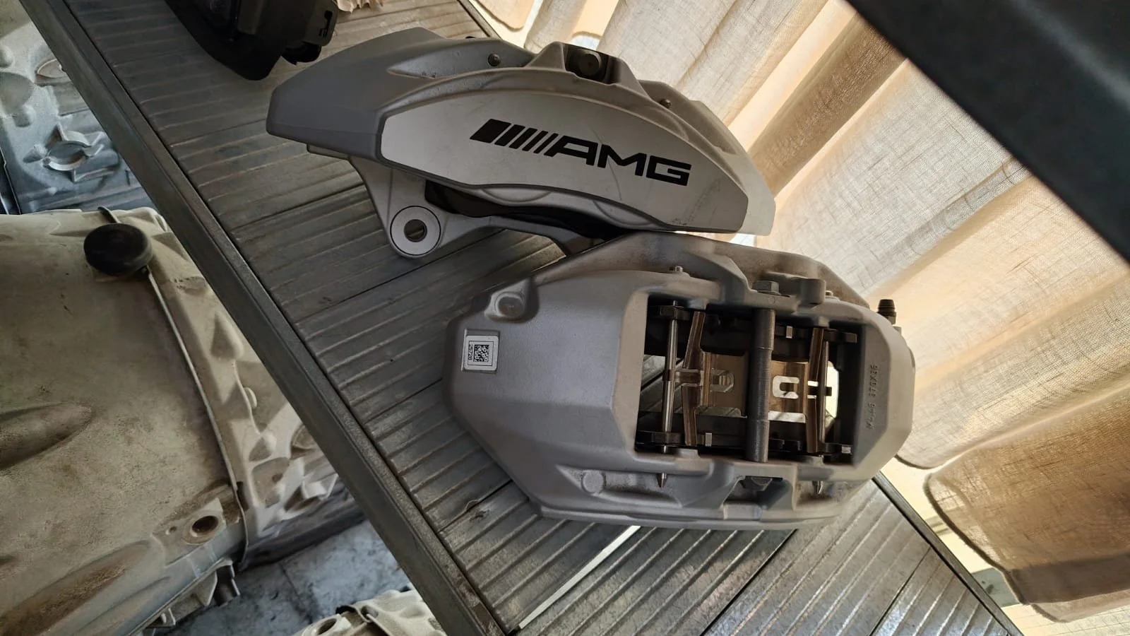 A silver AMG branded automotive brake caliper and brake housing with visible pistons, placed on a workbench in a workshop with curtains in the background.