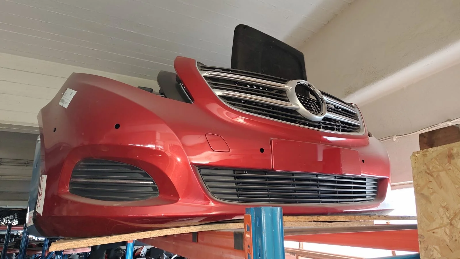 Red car front bumper with grille and headlights, stored on a wooden support in a workshop.