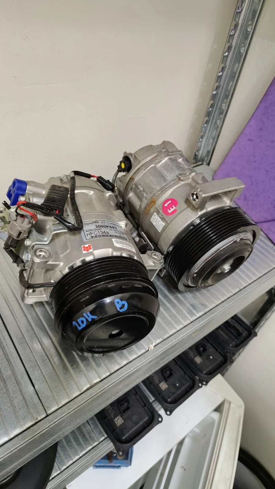 Two automotive air conditioning compressor units placed on a metallic surface in a workshop.