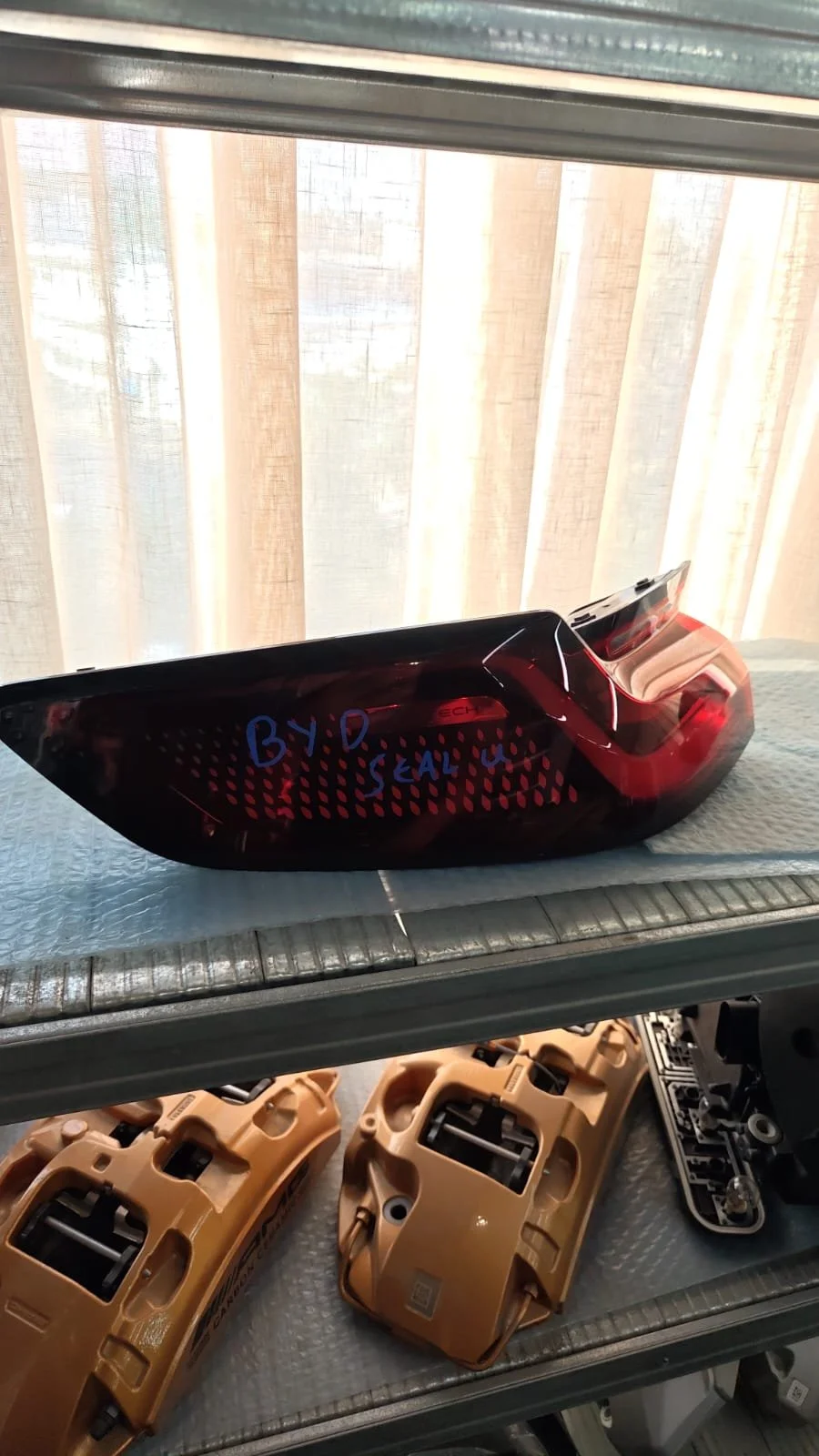 Rear taillight of a vehicle with a handwritten note 'BY: D Seal' on it, on a metal shelf with other vehicle parts below.