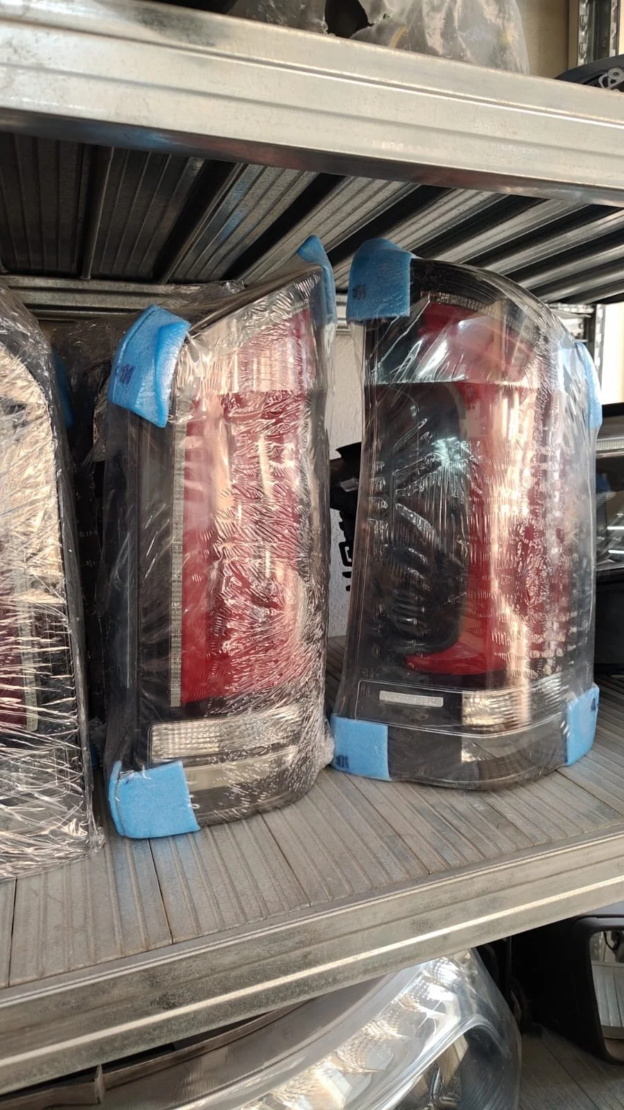 Two car tail lights wrapped in plastic and secured with blue painter's tape on a metal shelf.