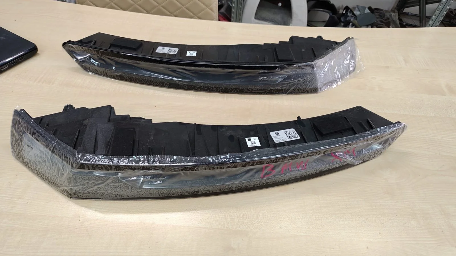 Two automotive headlight assemblies placed on a wooden table, wrapped partially in plastic and labeled with stickers and handwriting.