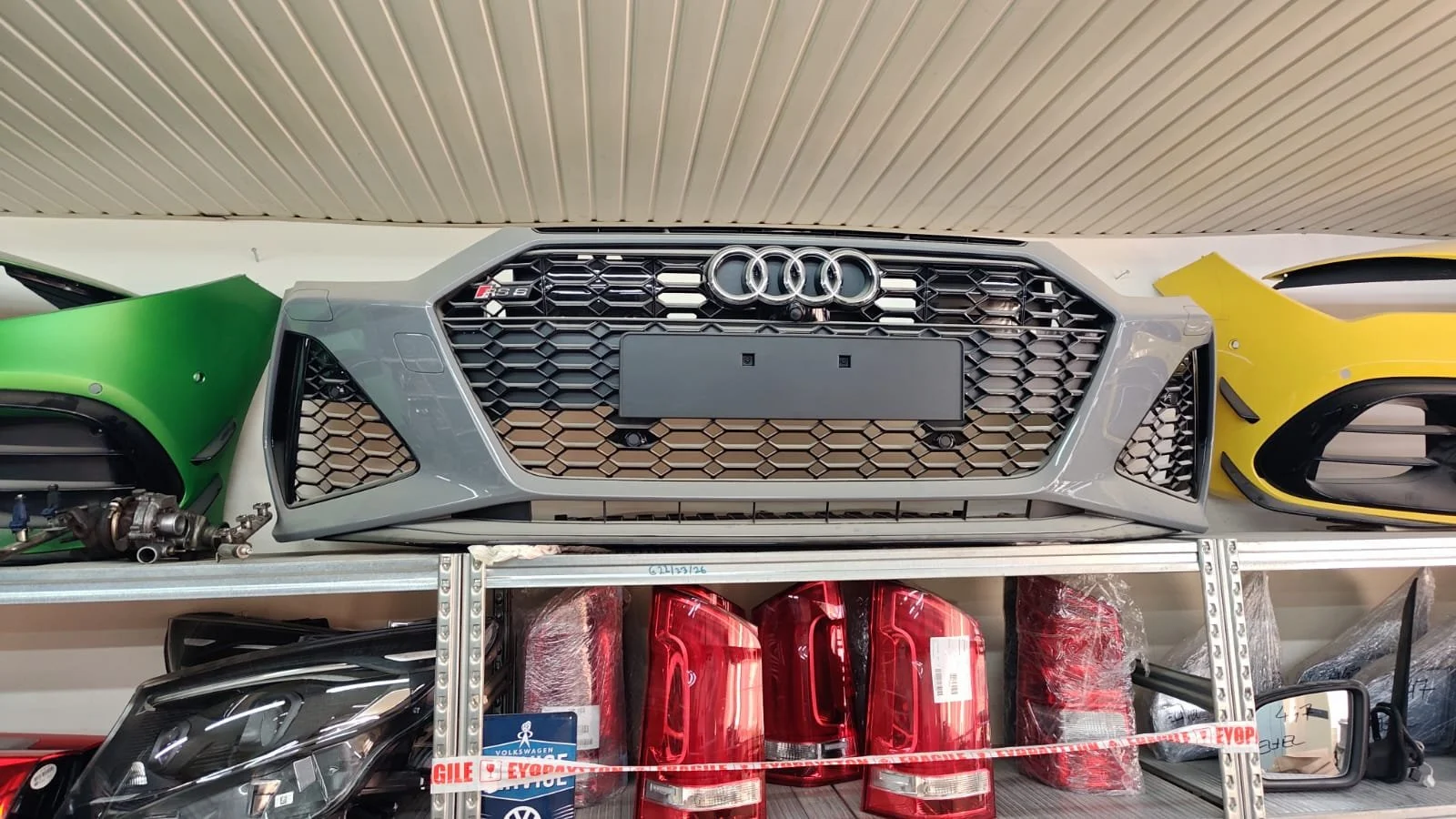 Car front grille with four rings logo, surrounded by green and yellow car parts, on a metal shelf with red and black tail lights underneath.