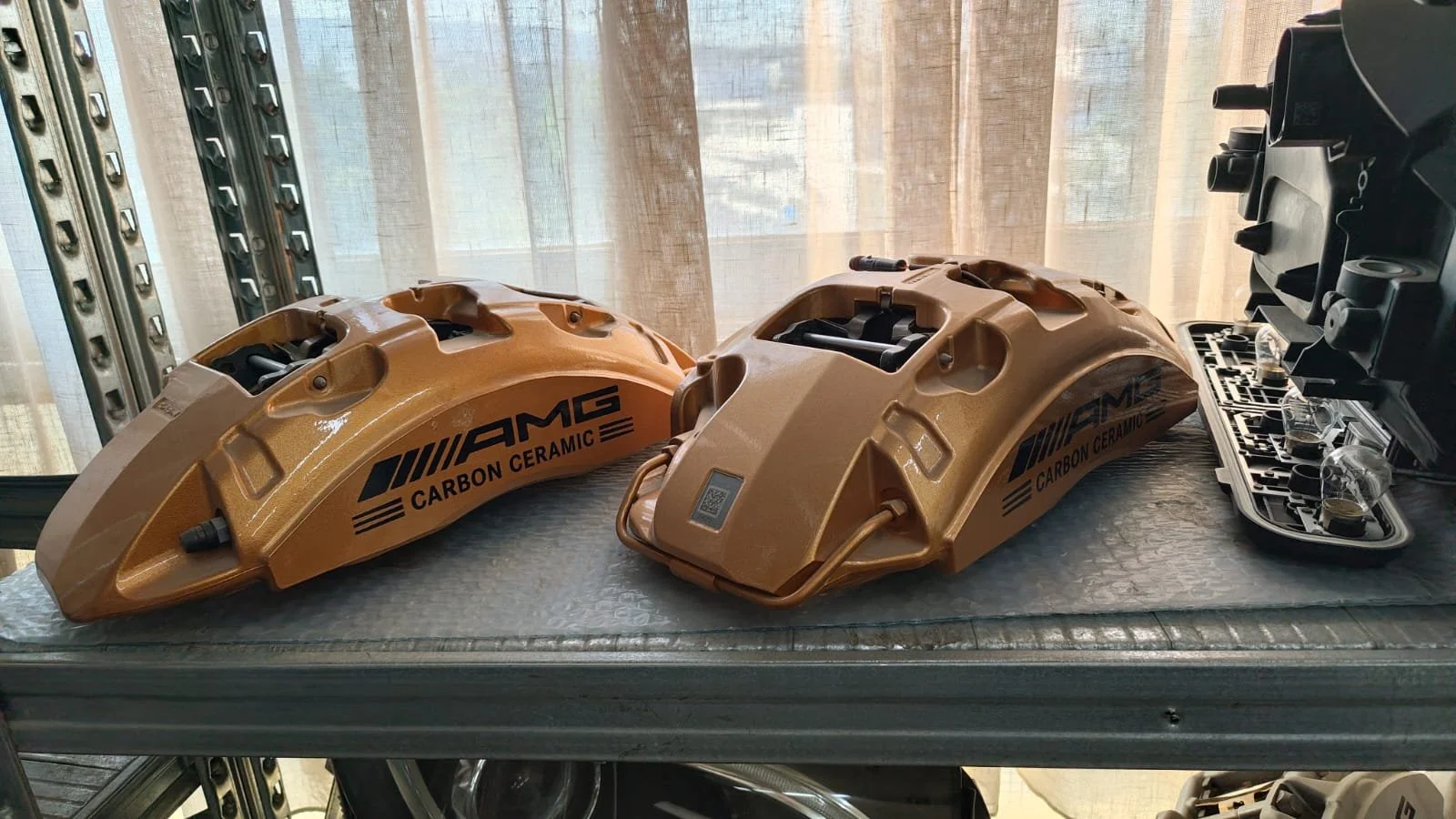 Two orange carbon ceramic brake calipers with AMG branding on a workshop table, with various tools and components nearby.