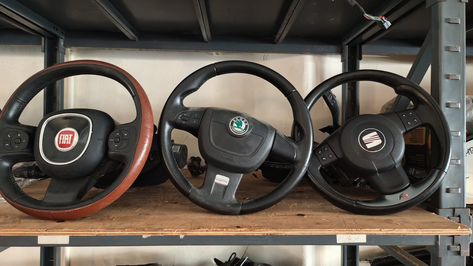 Three car steering wheels placed on a wooden shelf, with a Fiat on the left, a Škoda in the middle, and a SEAT on the right.