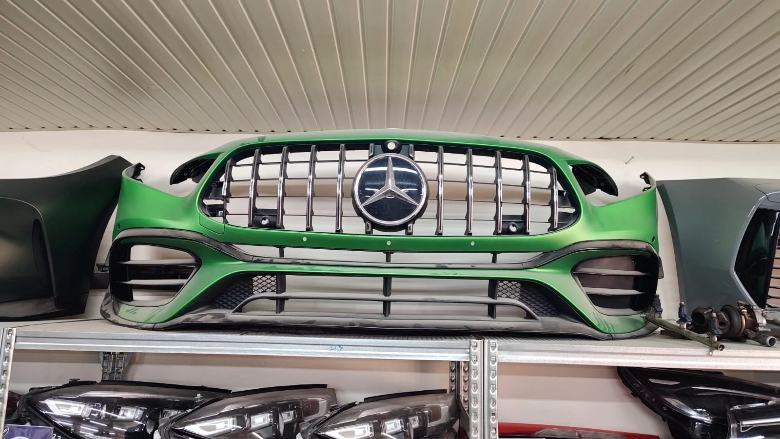 Green and black Mercedes-Benz car front grille with emblem, placed on a shelf in a garage or workshop.