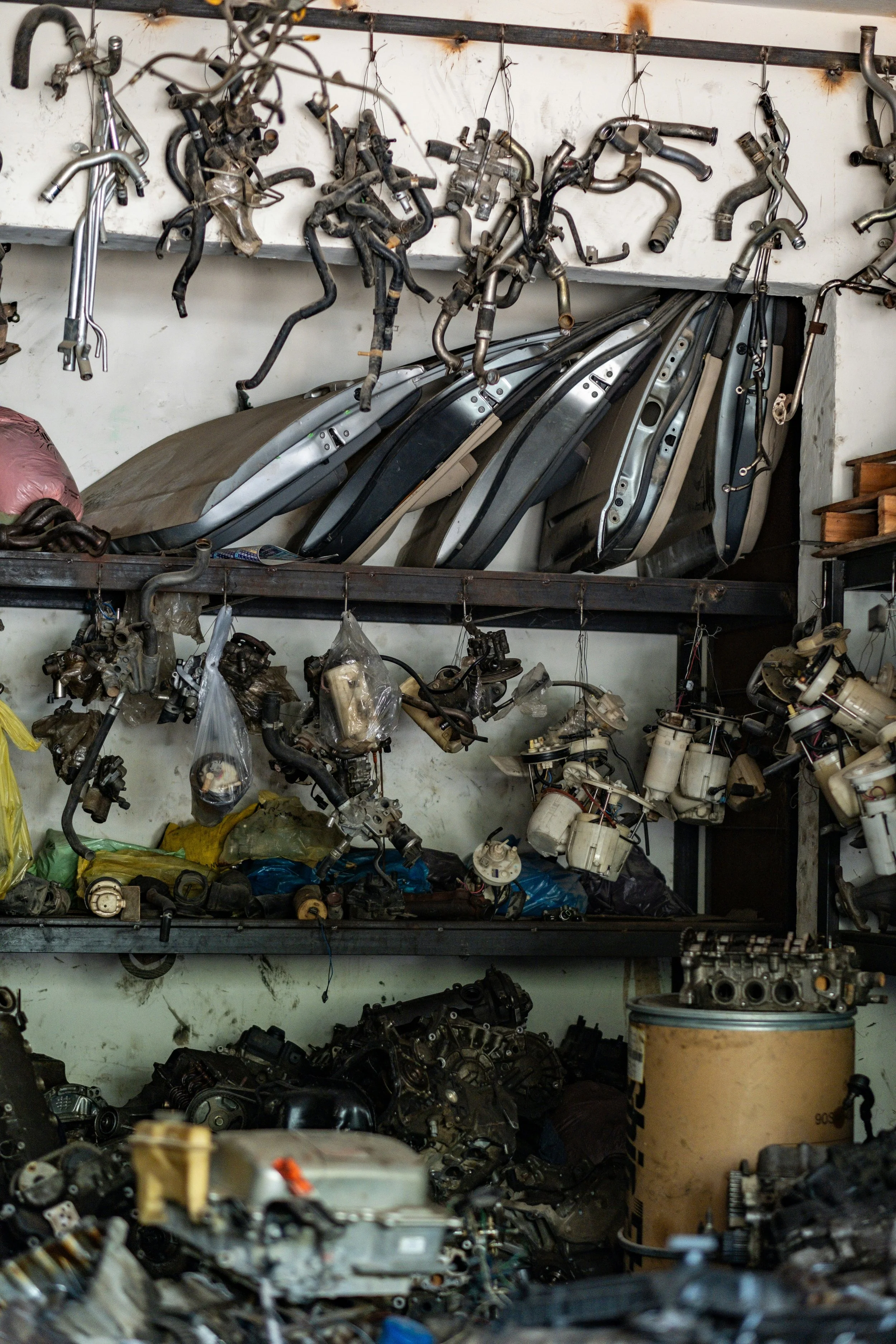 A cluttered workshop with shelves holding disassembled car engine parts, hoses, and body panels, some hanging from hooks or lying on the shelves.
