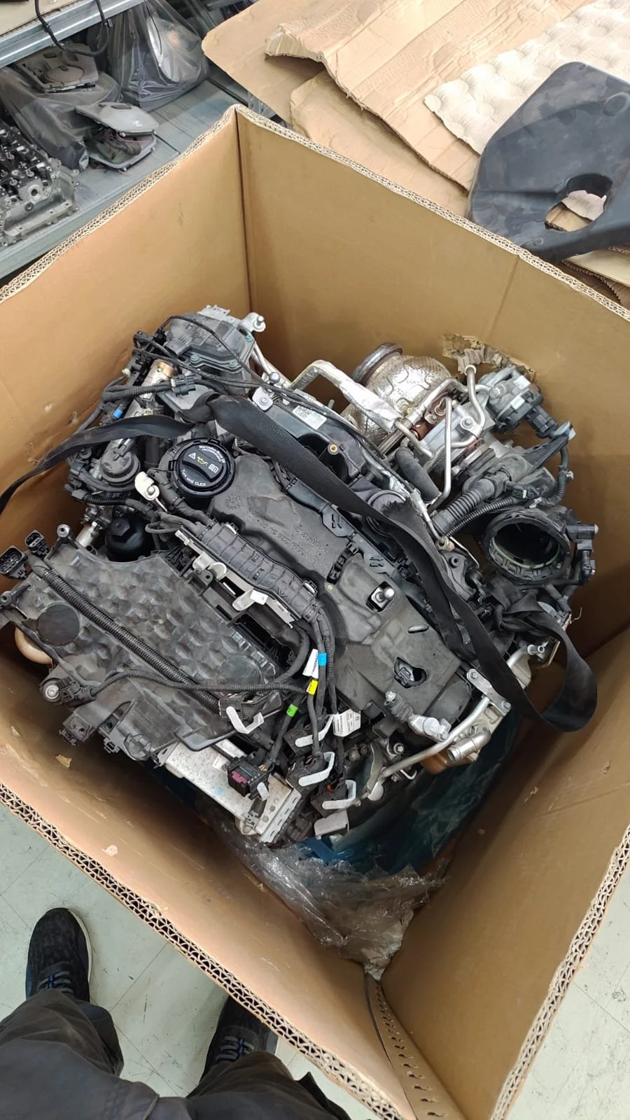 An engine inside a large cardboard box on a workbench, with various engine parts and tools nearby.