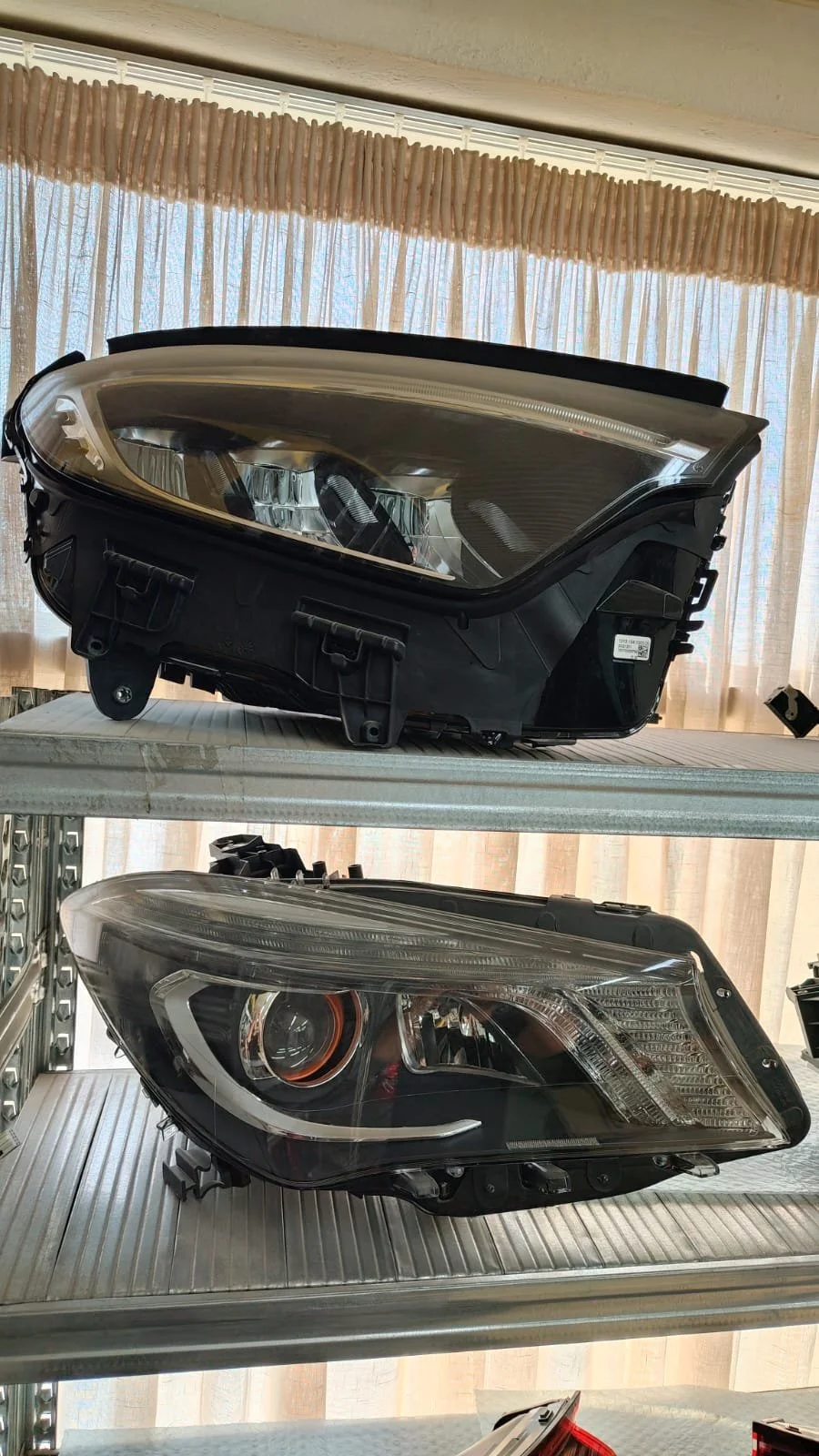 Two car headlights, one larger and one smaller, placed on a metal shelf with curtains in the background.