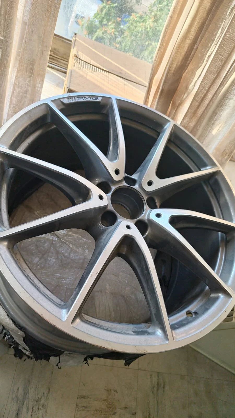 A close-up of a silver, multi-spoke alloy wheel rim for a car, placed near a window with beige curtains and some greenery visible outside.