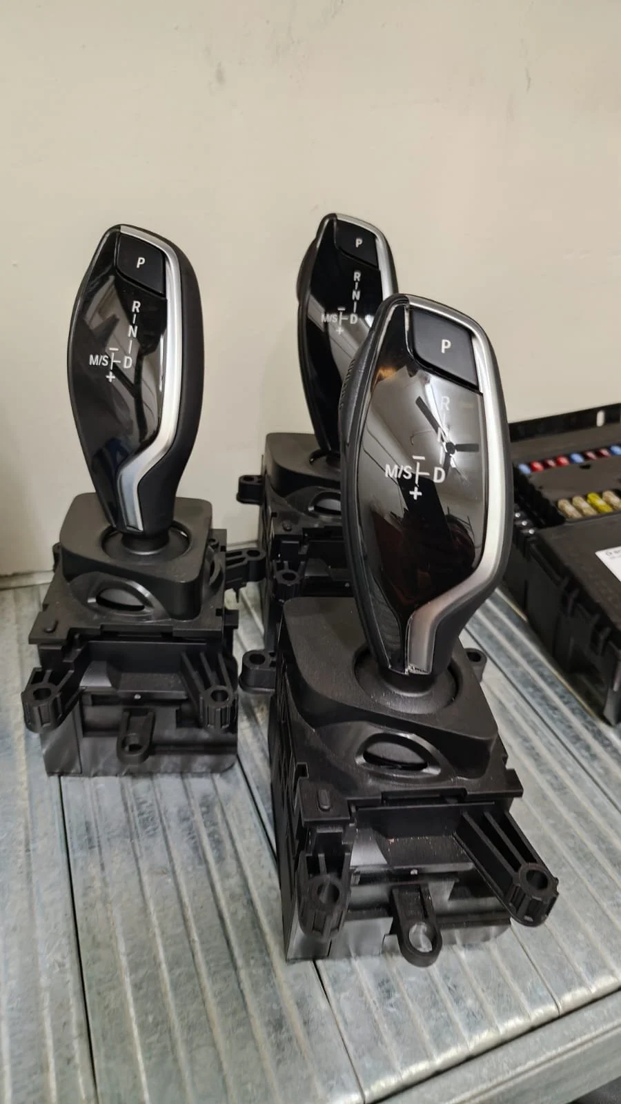 Three car gear shifters with digital displays, mounted on black bases, placed on a metallic surface, with a set of assorted electronic components in the background.