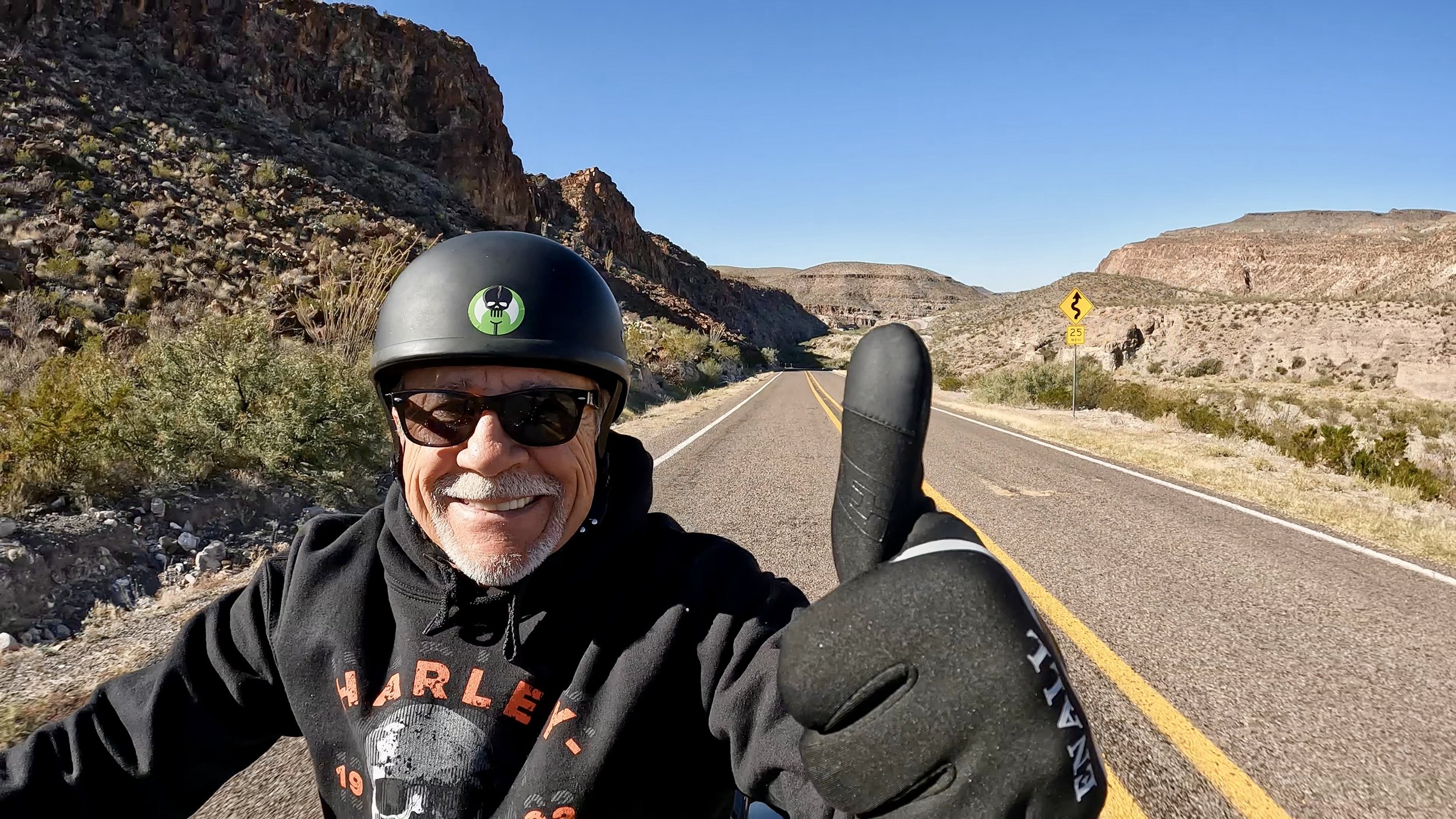 A smiling man in motorcycle gear taking a selfie on a desert highway with rocky formations and a winding road in the background.