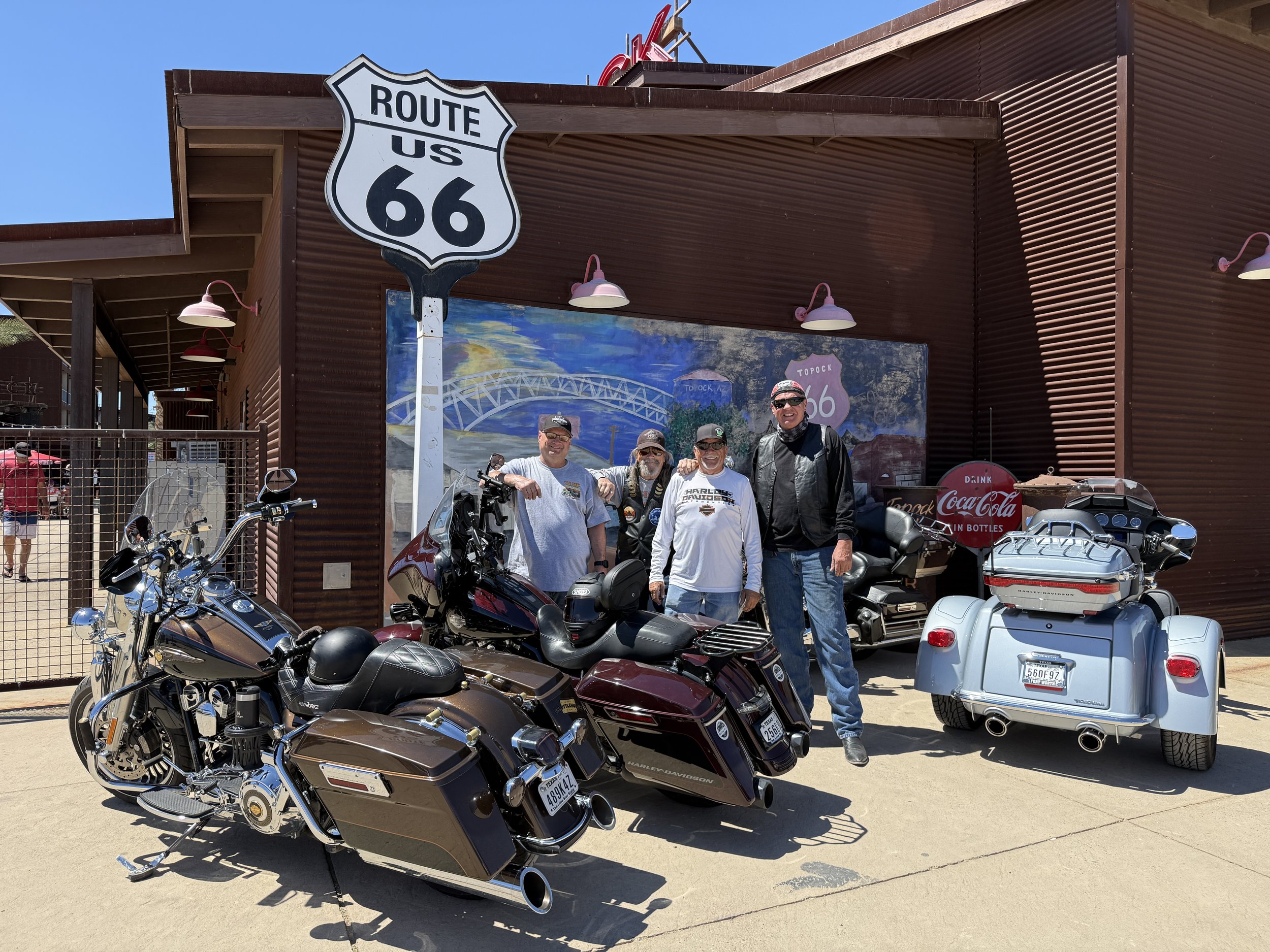 Four men standing in front of parked motorcycles and cars outside a building with a Route 66 sign and a painted mural