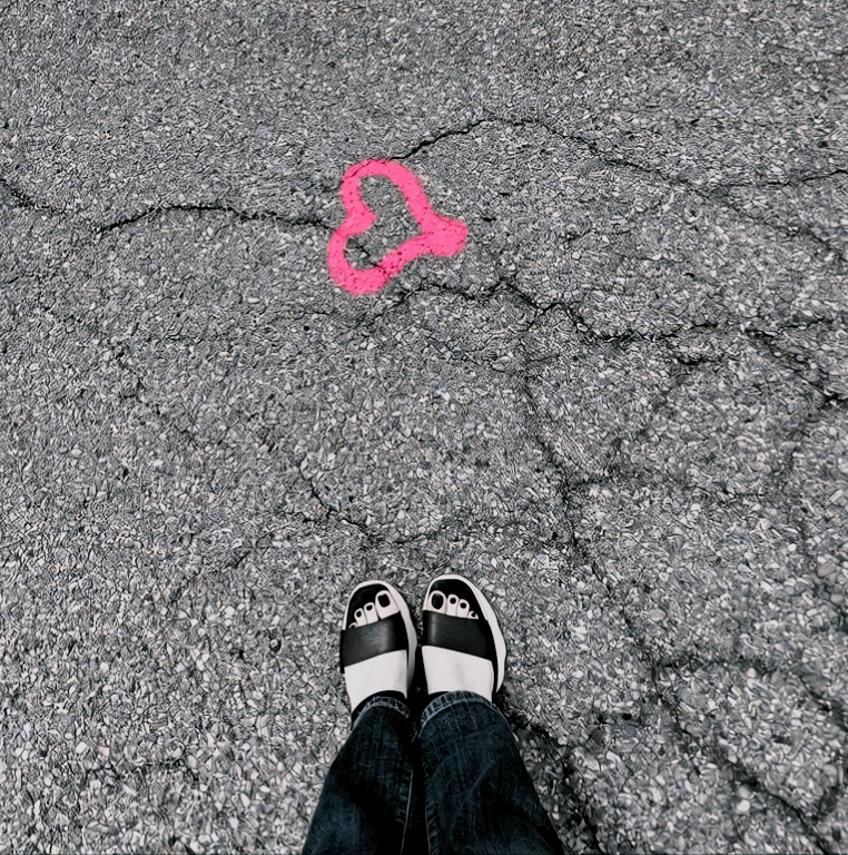View of a person's feet in black and white sandals on a cracked asphalt surface with a pink spray-painted heart outline on the ground in front of them.