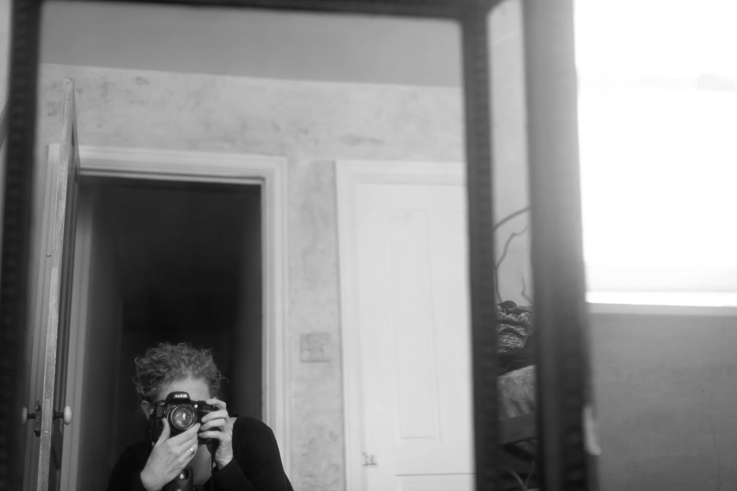 A person with curly hair holding a camera taking a photo in front of a mirror in a room with doorways and a window.