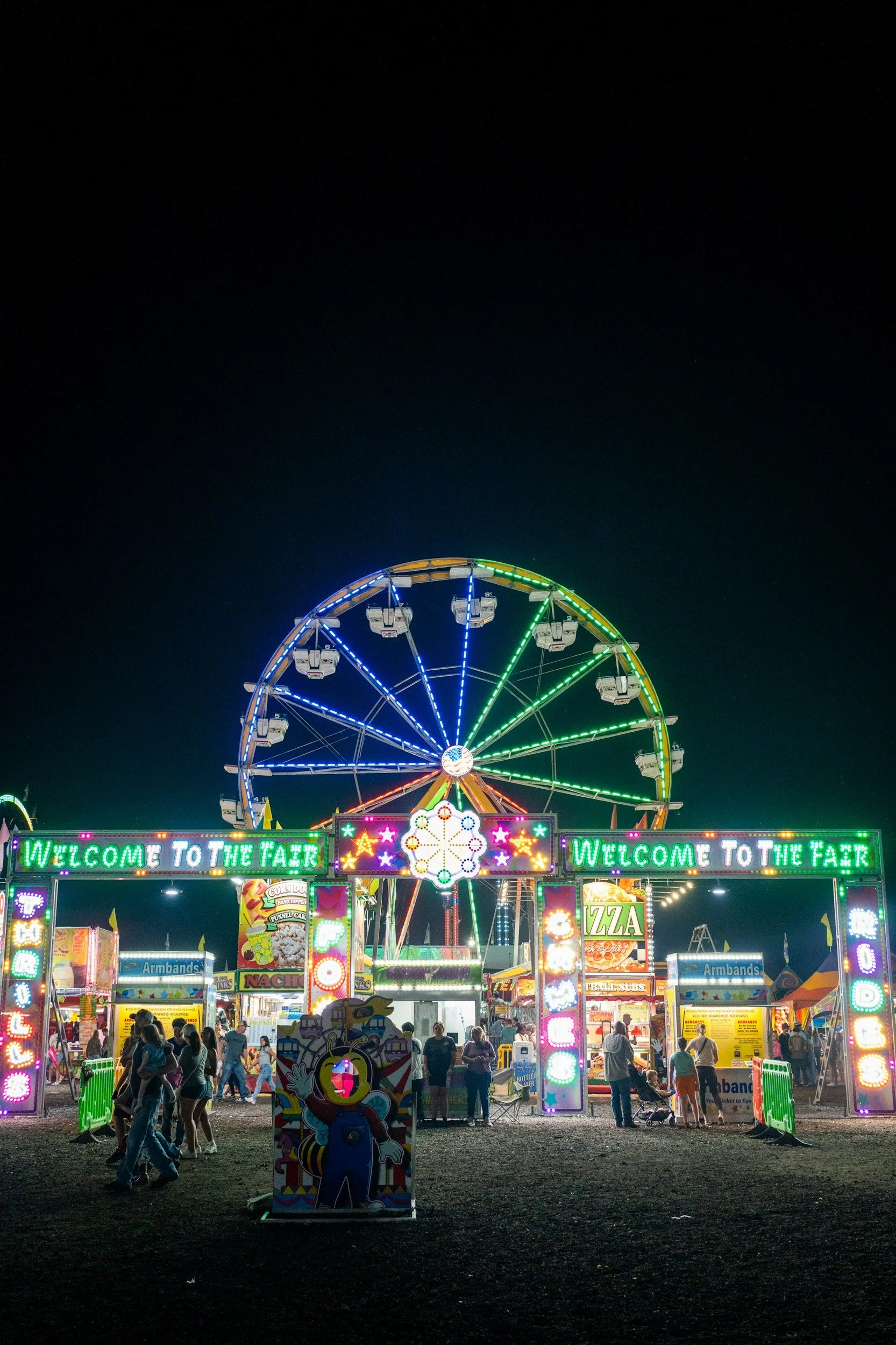Nighttime scene at a fair with a large Ferris wheel lit with colorful lights, a carnival entrance with bright neon signs that say 'Welcome to the Fair,' and various food and game booths with people walking around.