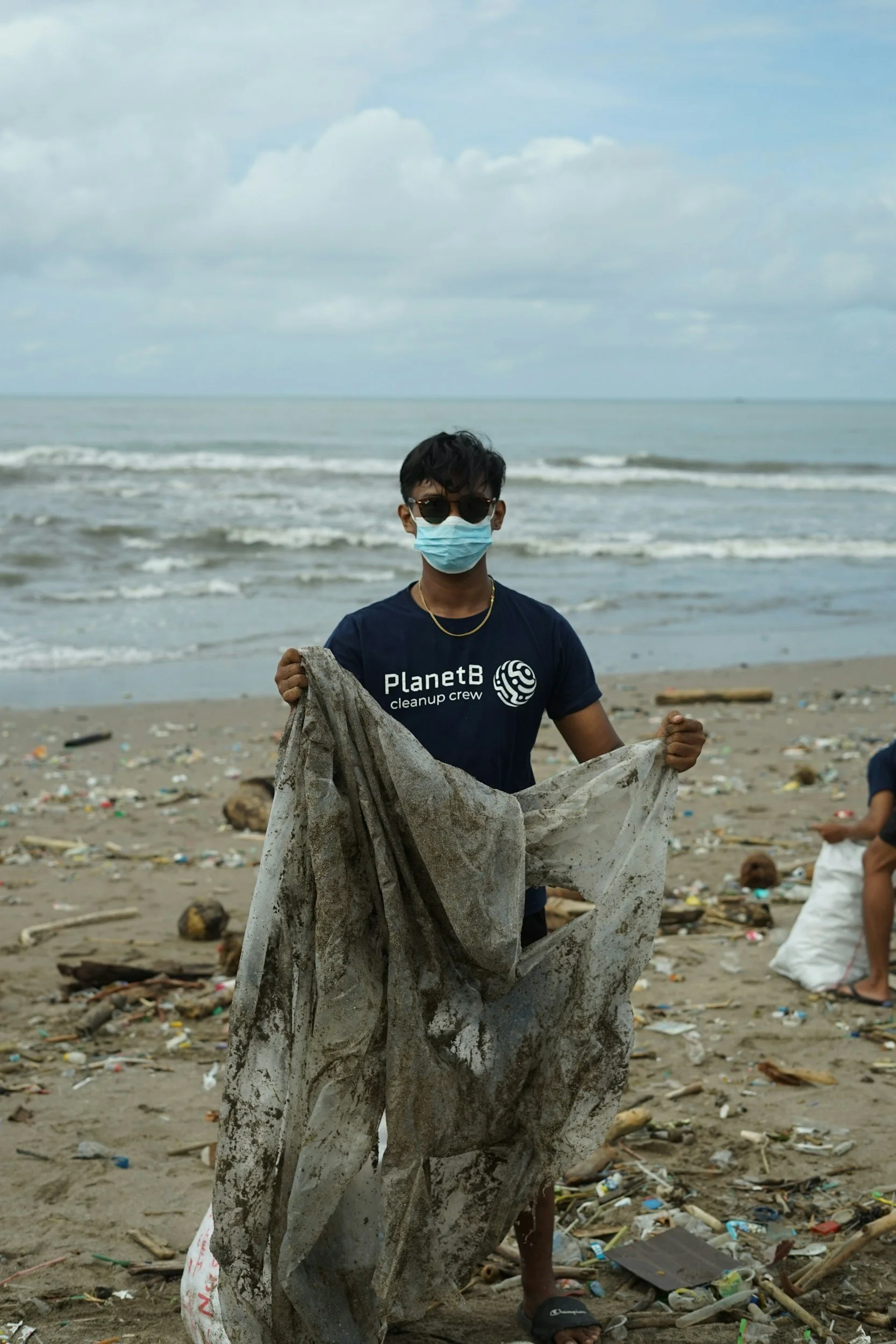 A person wearing sunglasses and a face mask on a polluted beach, holding a large dirty plastic bag amid trash and debris with the ocean and cloudy sky in the background.