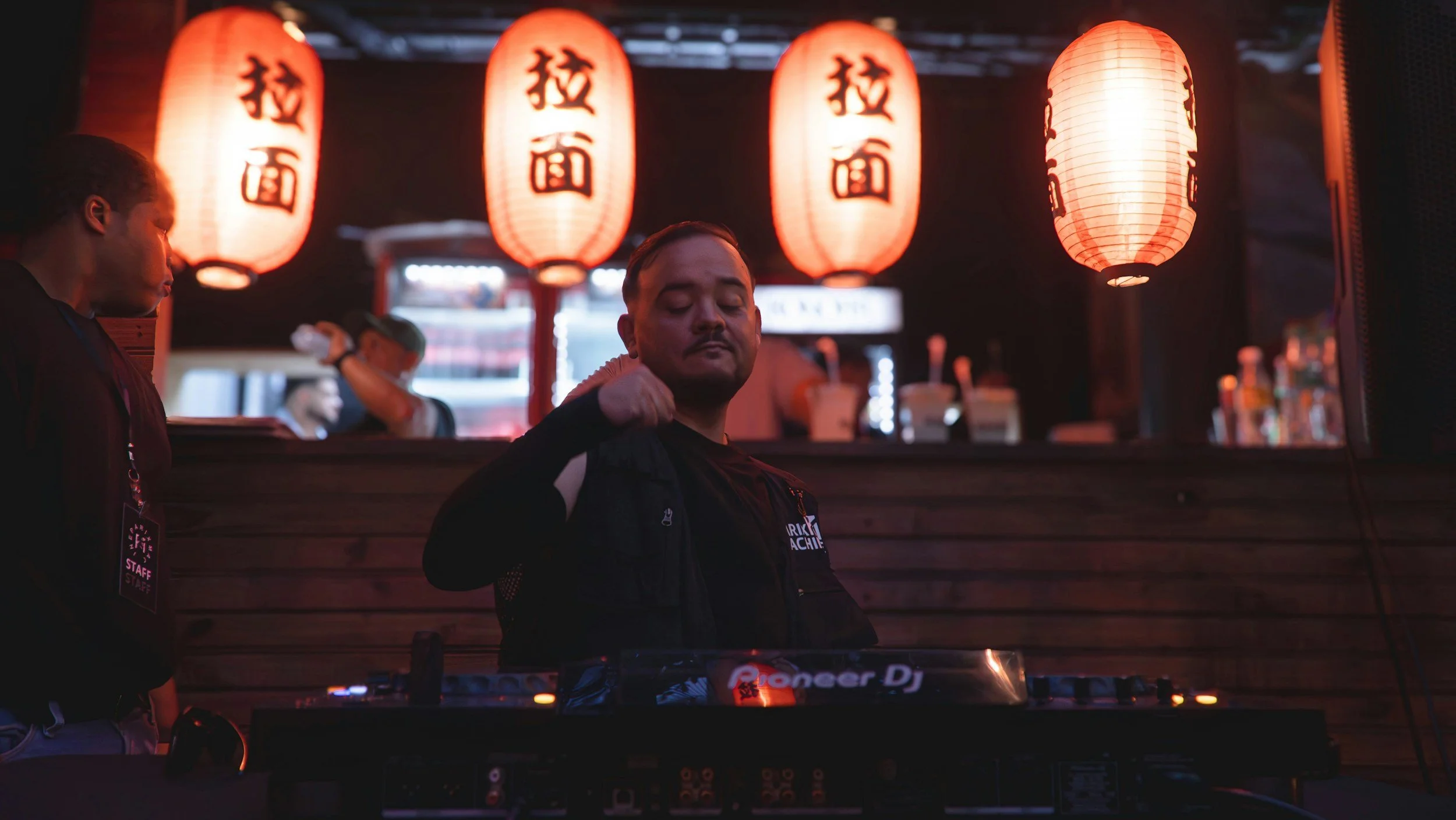 A man with closed eyes standing behind a DJ with Pioneer DJ equipment, illuminated by five red paper lanterns with black Chinese characters hanging above them in a dimly lit indoor setting.