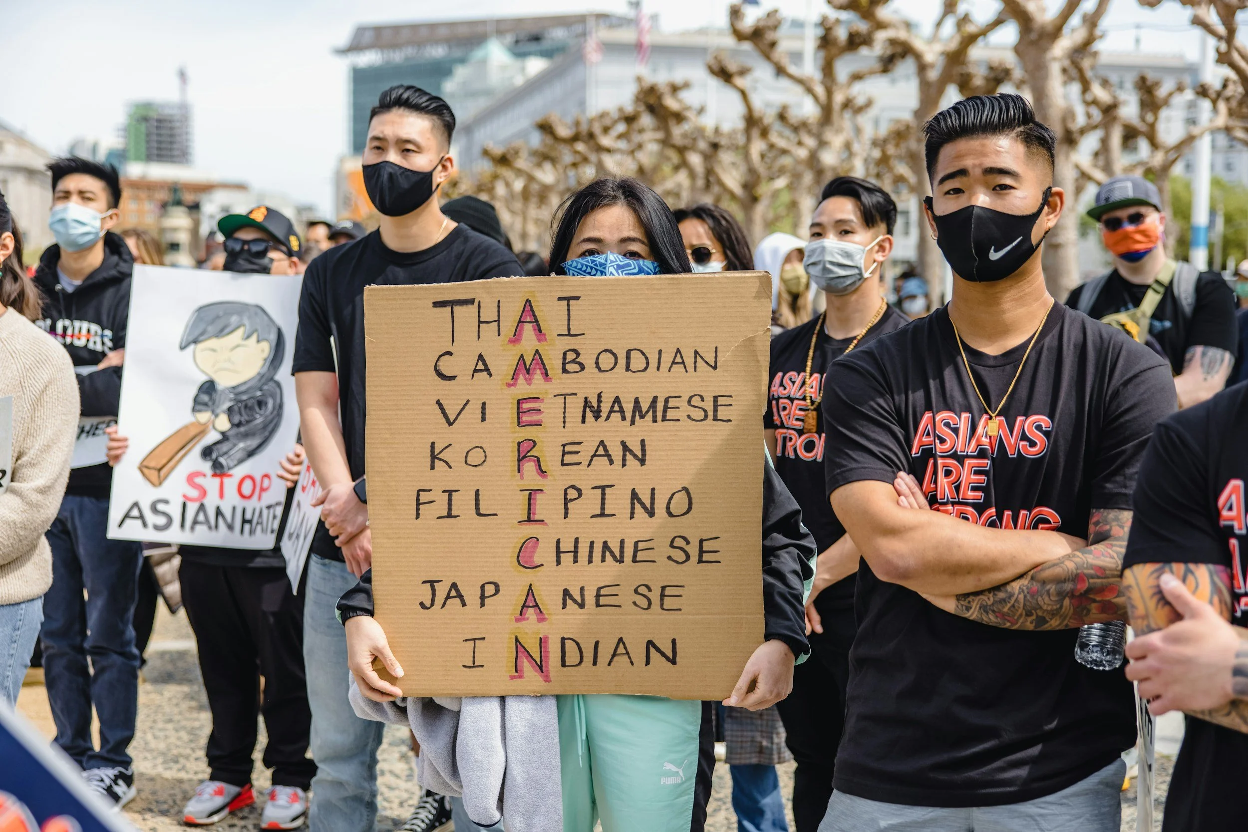 A group of people protesting outdoors, wearing masks, holding signs, with a woman in the center holding a cardboard sign listing Asian countries and the word 'Asian' vertically.