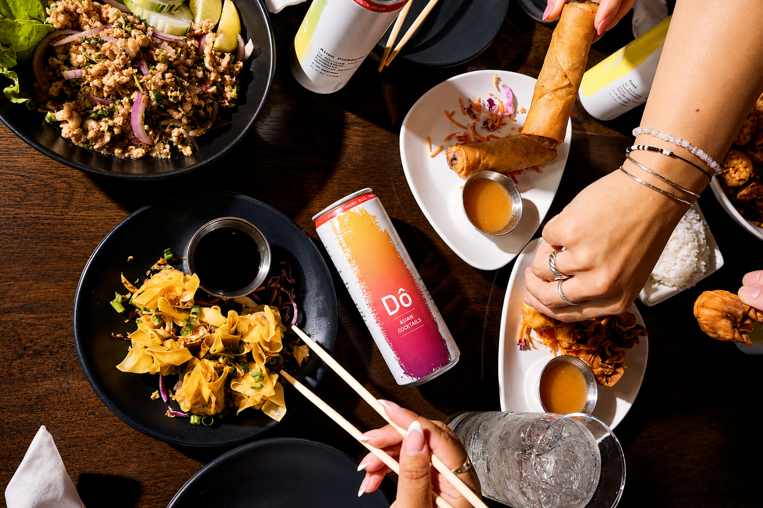 Top-down view of a table with various Asian dishes, including spring rolls, dumplings, a rice and peanut salad, and drinks, including canned Do Asian cocktails, a glass of water, and soy sauce in small bowls.