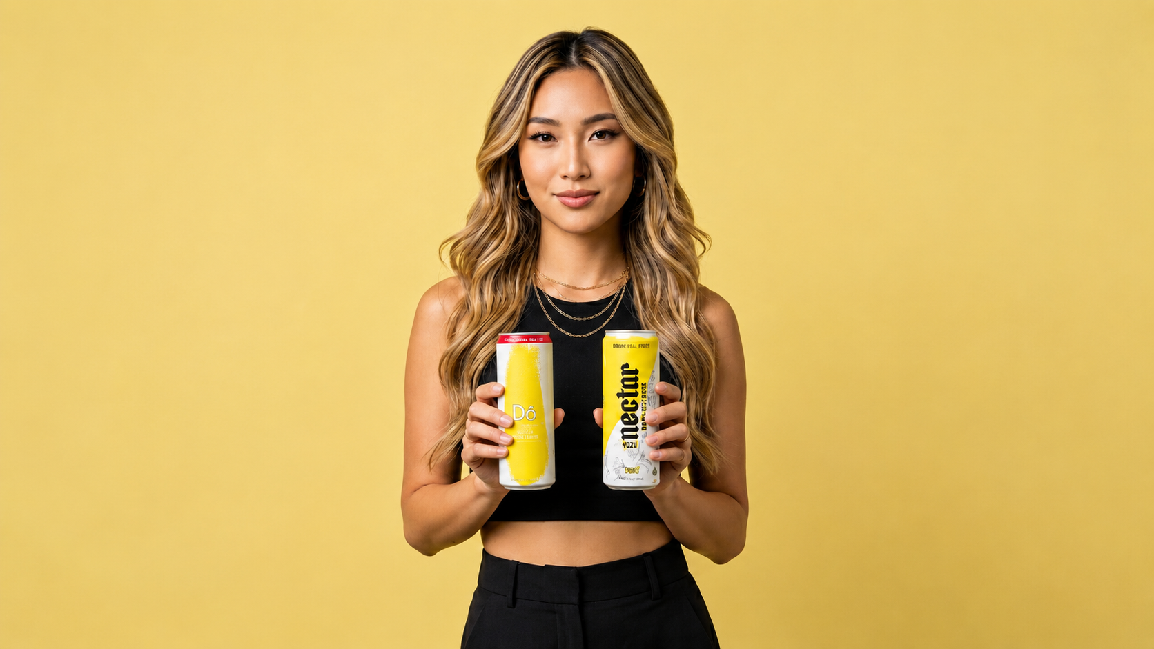 An Asian Baby Girl (ABG) with long wavy blonde hair, wearing a black crop top and black pants, holding two cans of Dô Asian Cocktails against a yellow background.