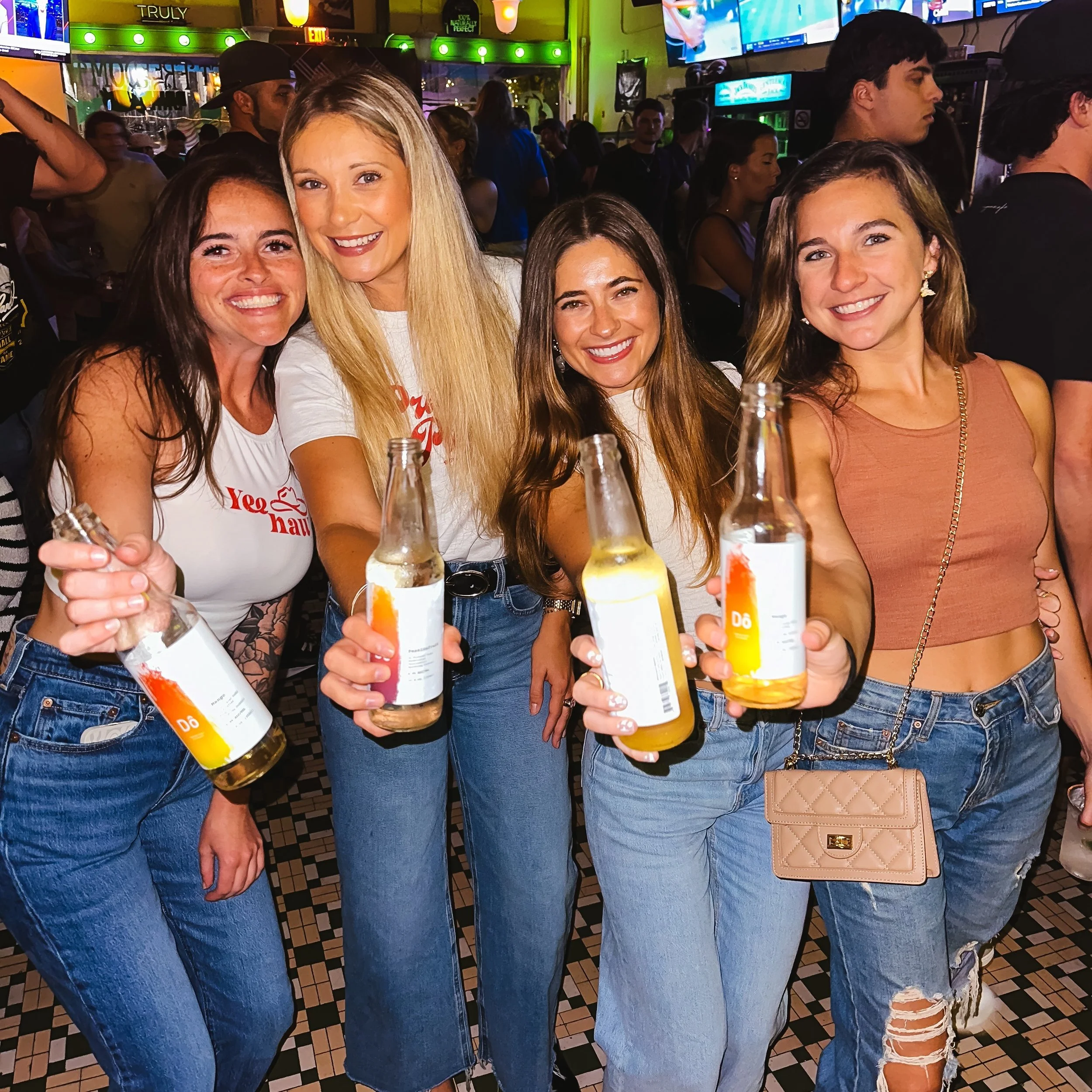 Four women dressed in casual jeans and tops holding Do Asian Cocktails in a lively, crowded bar or restaurant. The background features multiple television screens and neon signs.