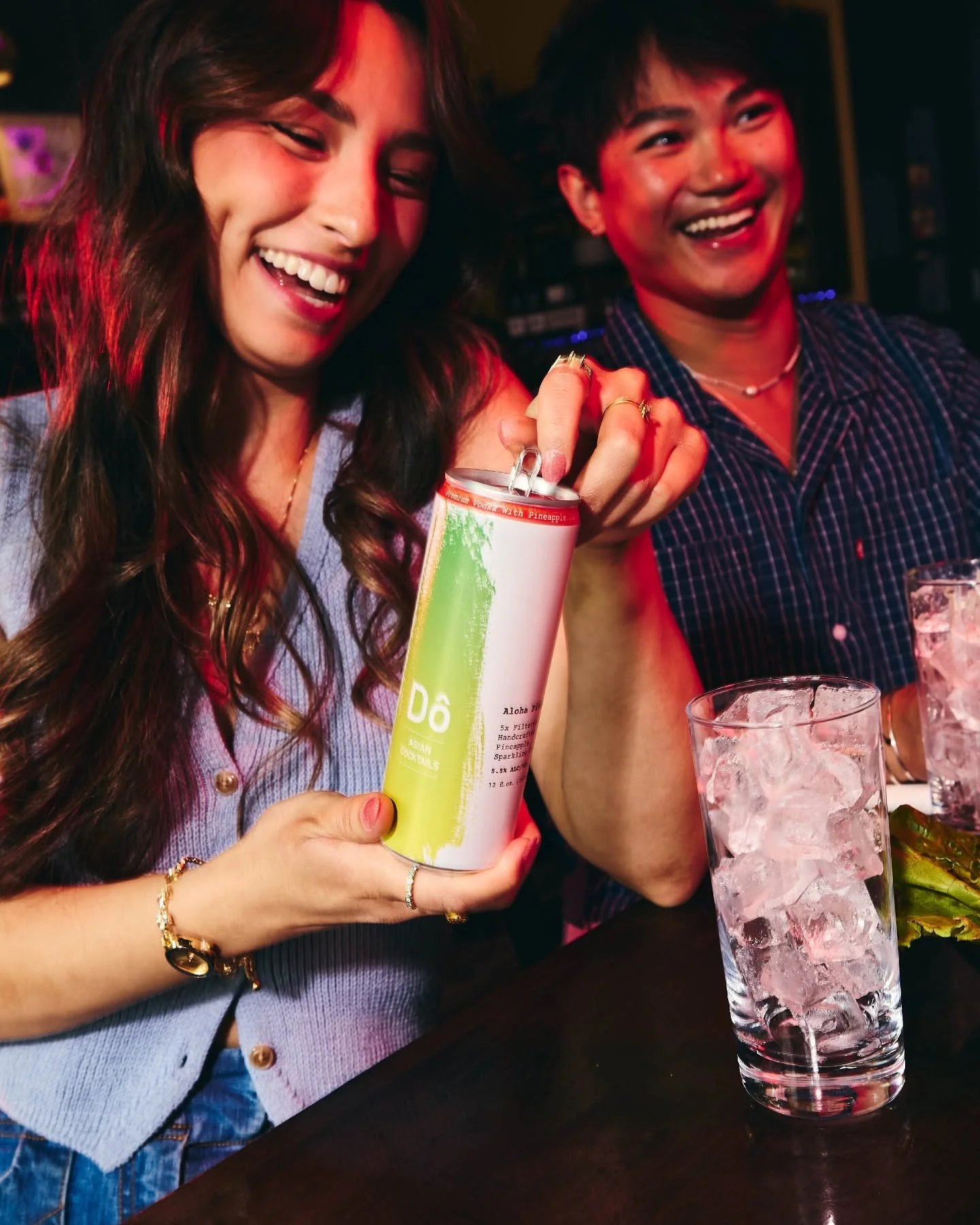 Two young women smiling and enjoying drinks at a social gathering, one holding a Do Asian Cocktail Fruit Vodka Seltzer, and the other with a glass of ice Do Asian Cocktail Fruit Vodka Seltzer.