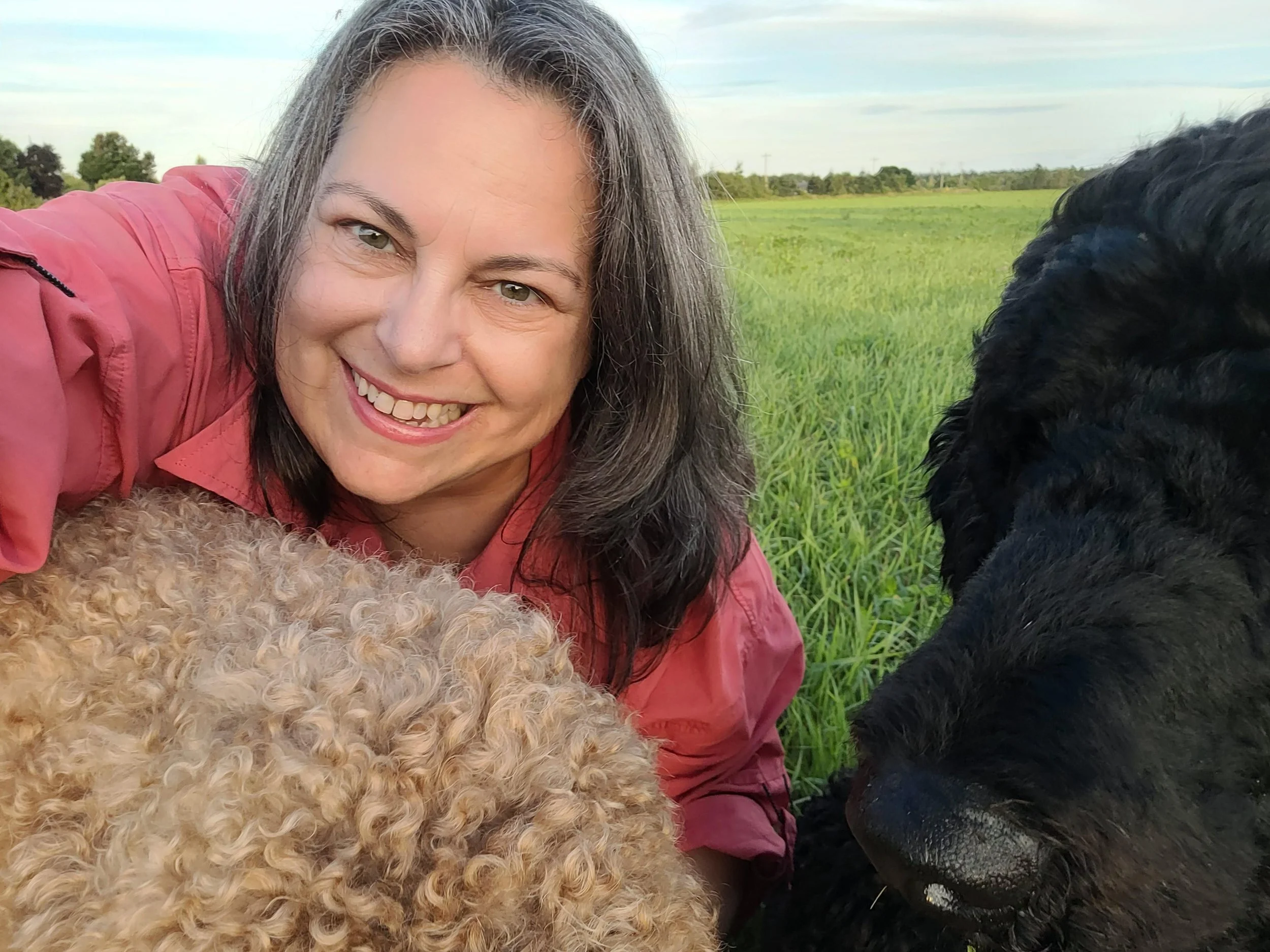 Smiling woman with gray hair wearing a pink jacket, lying on the grass next to a black dog in a green field with trees and blue sky in the background.