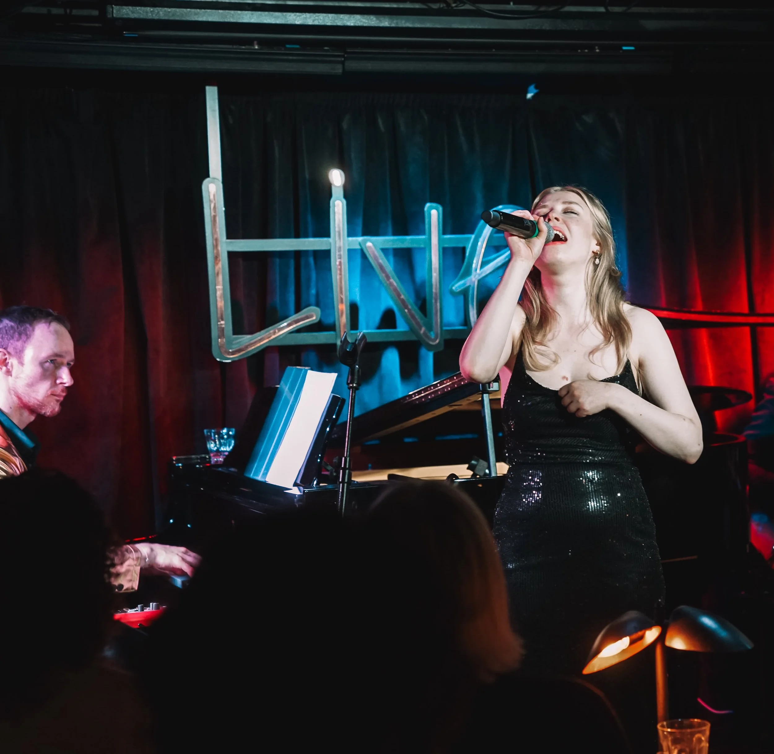 A woman singing into a microphone during a live performance, with a piano in front of her and a dark curtain backdrop.