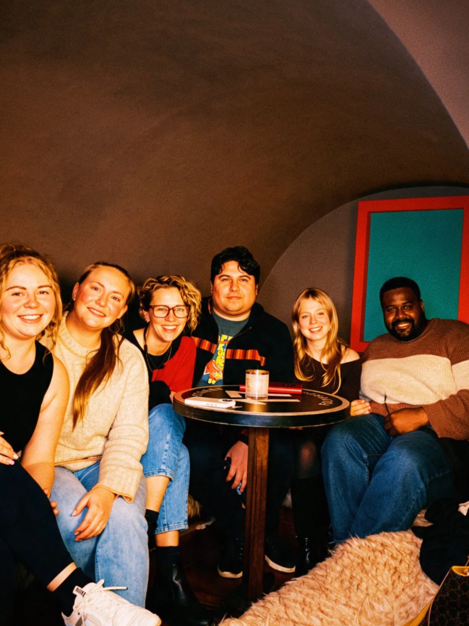 Group of six friends sitting around a table in a cozy indoor setting, smiling at the camera.