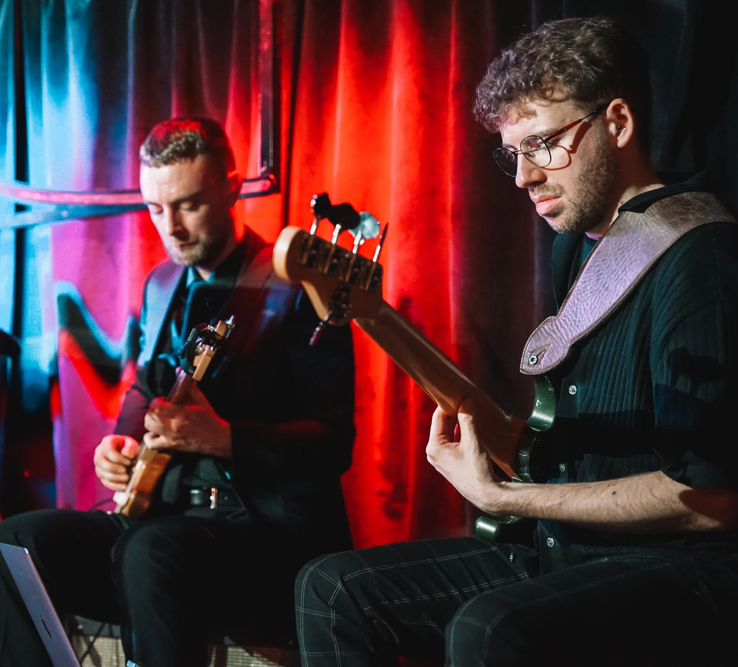 Two musicians performing on stage near a red curtain; the musician on the right is wearing glasses and a dark shirt, playing an bass guitar, while the other musician on the left has short hair and facial hair, playing a guitar.