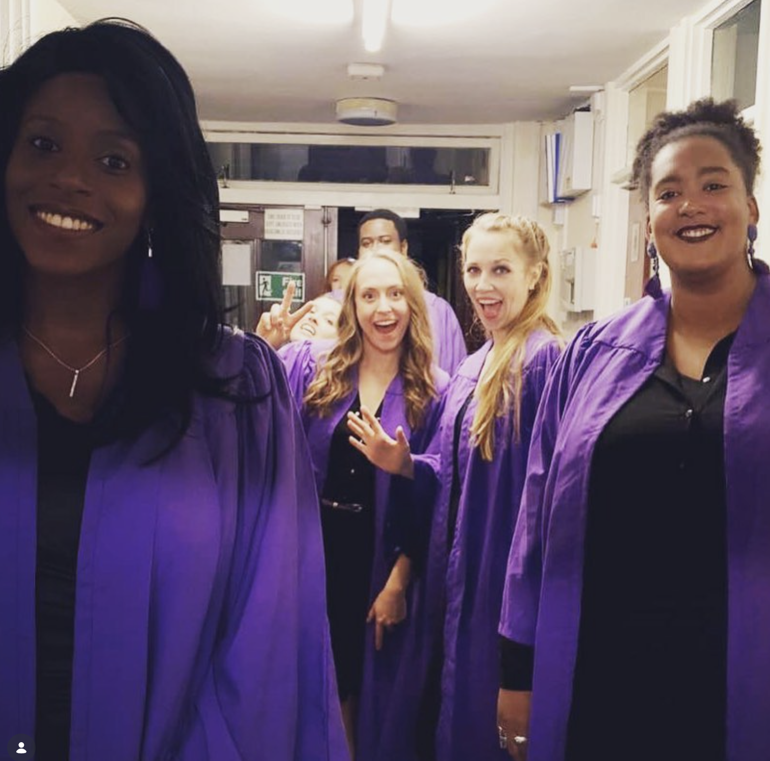Group of diverse young women wearing purple graduation gowns, celebrating and smiling indoors.