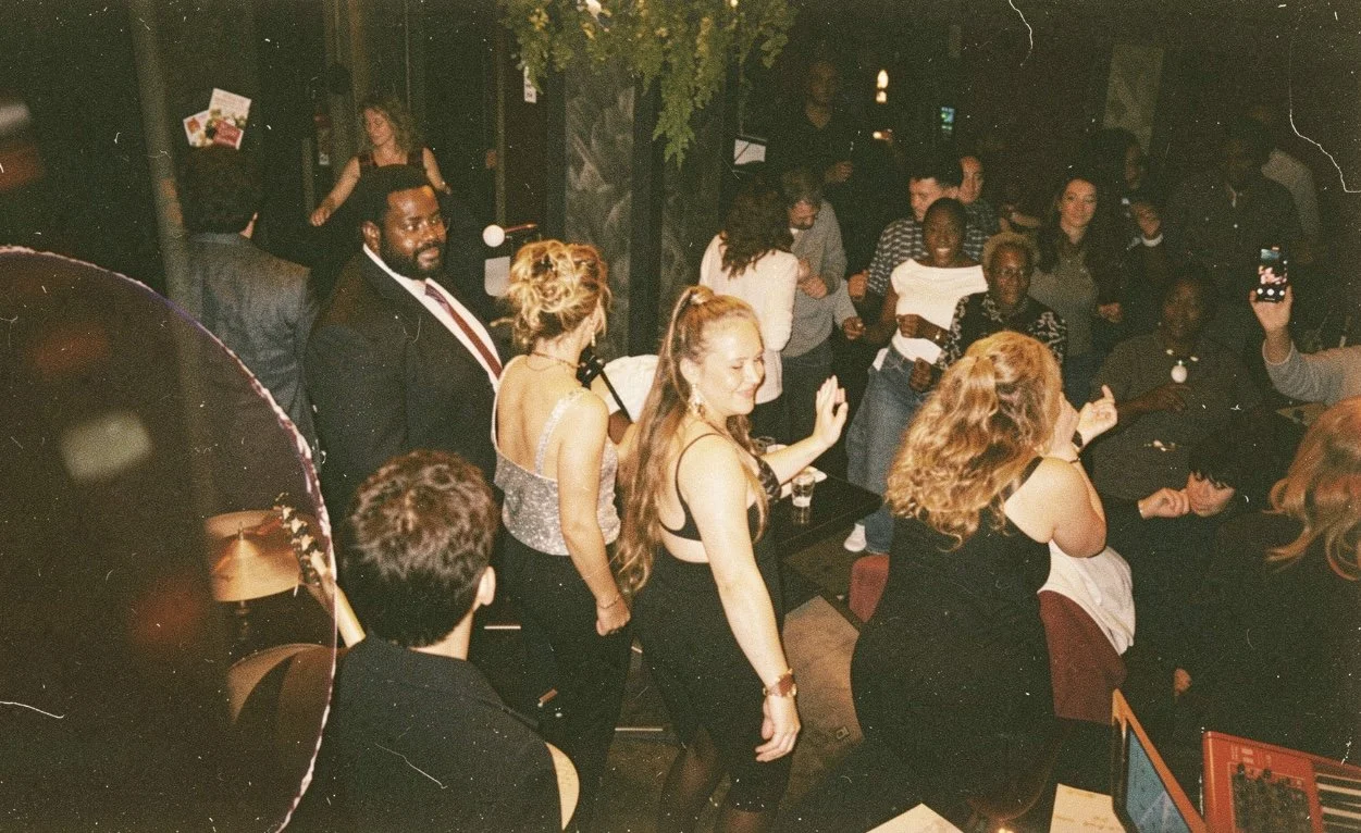People dancing and enjoying a party or celebration in a dimly lit indoor venue with a variety of guests, some taking photos, and a DJ or music setup.