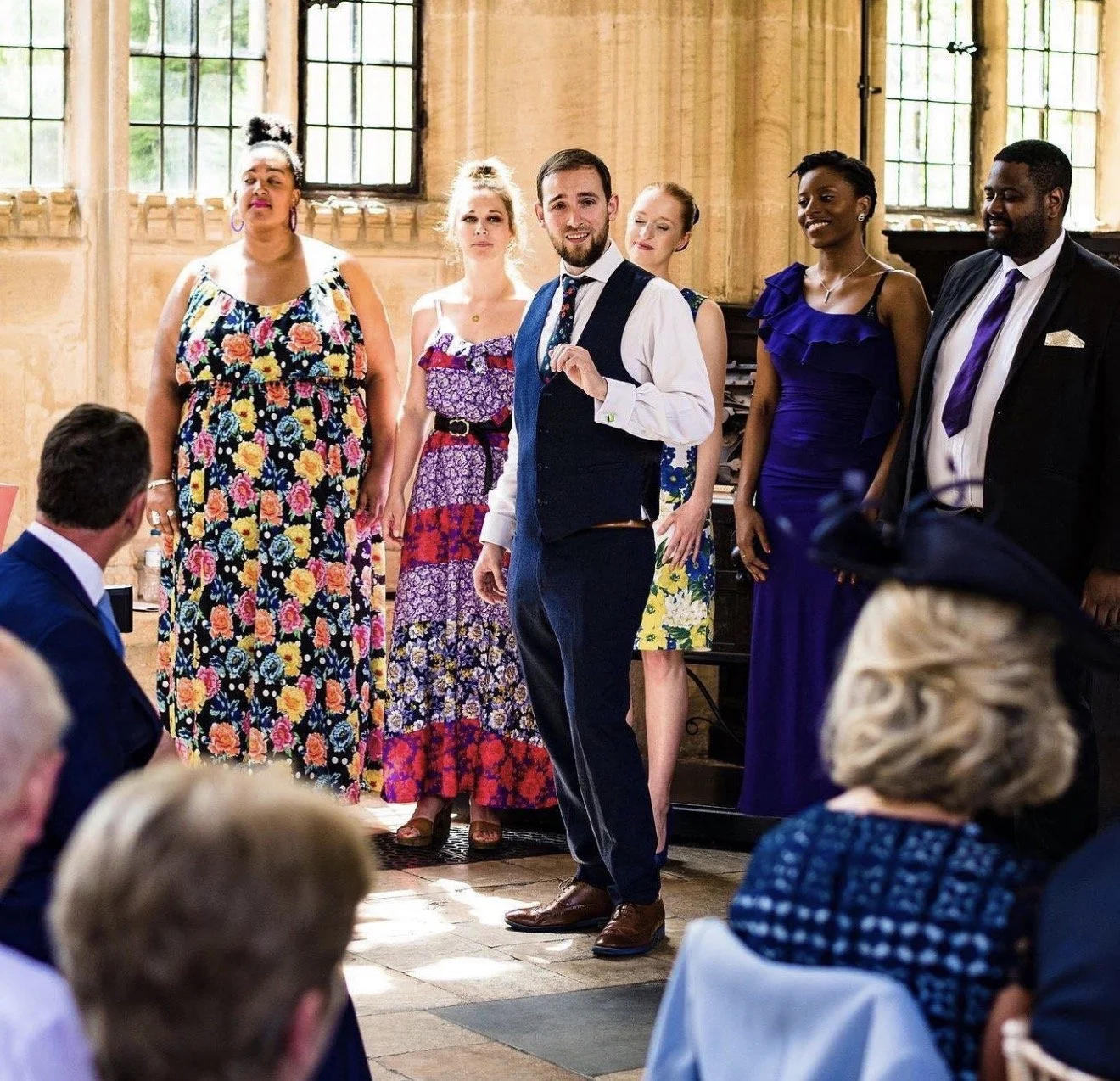 People standing in a room with high windows, dressed in colorful formal attire, with some seated at tables, during a social event or celebration.