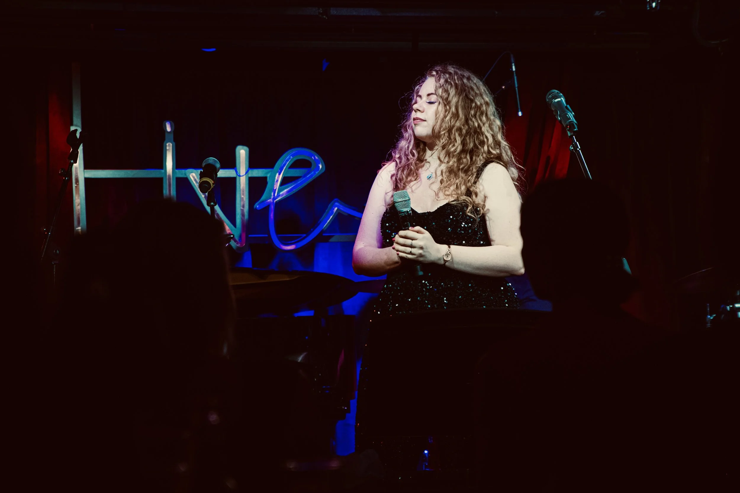 A woman with curly hair standing on stage holding a microphone, with a neon sign behind her that reads 'Live' and a red curtain on the side.