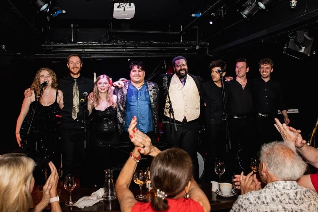 Musicians standing on stage after a performance, holding each other with an audience clapping and raising hands in appreciation.
