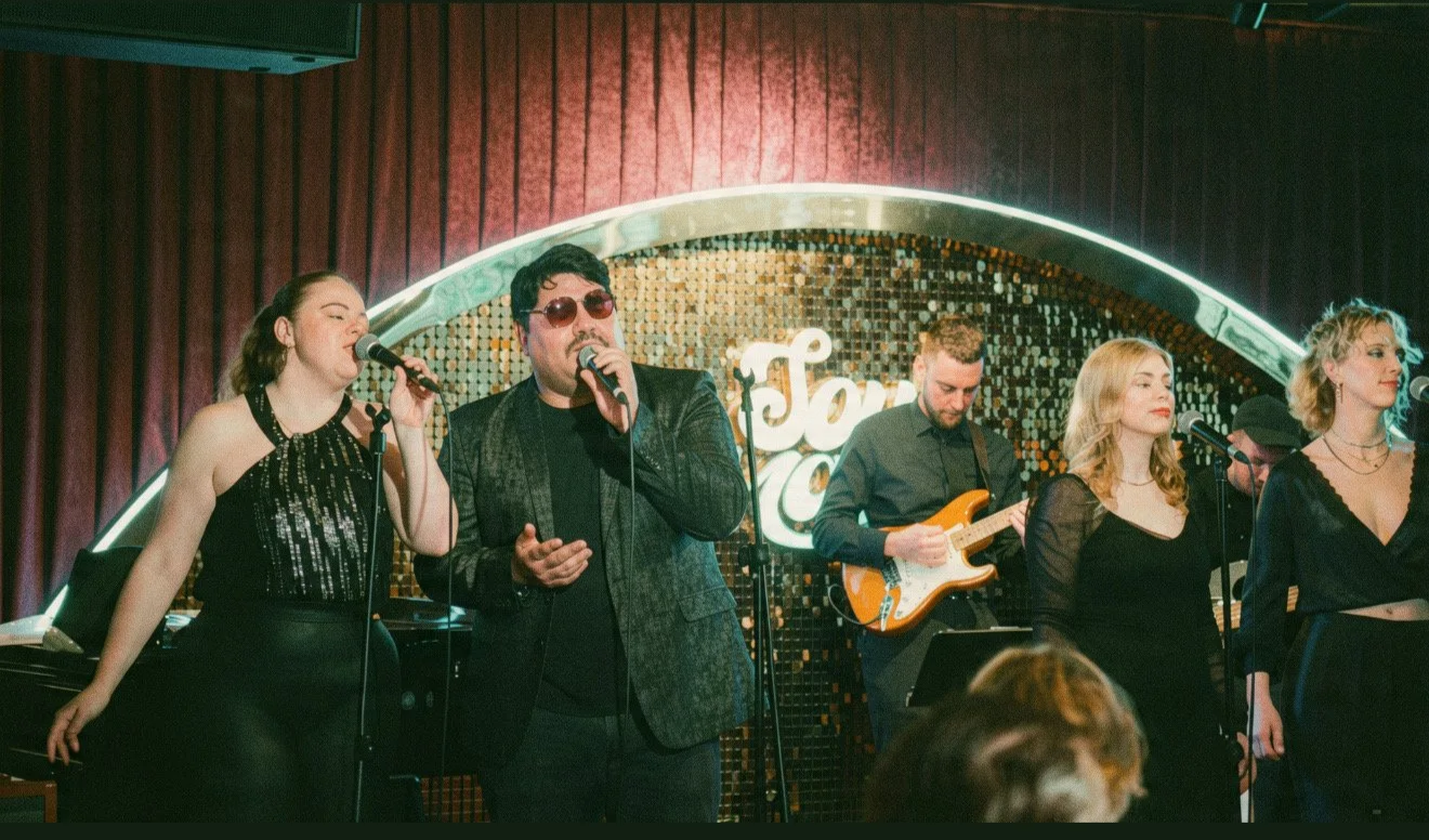 A group of six performers on stage at a jazz club, singing and playing music with microphones and an electric guitar, with a glowing sign and red curtains in the background.