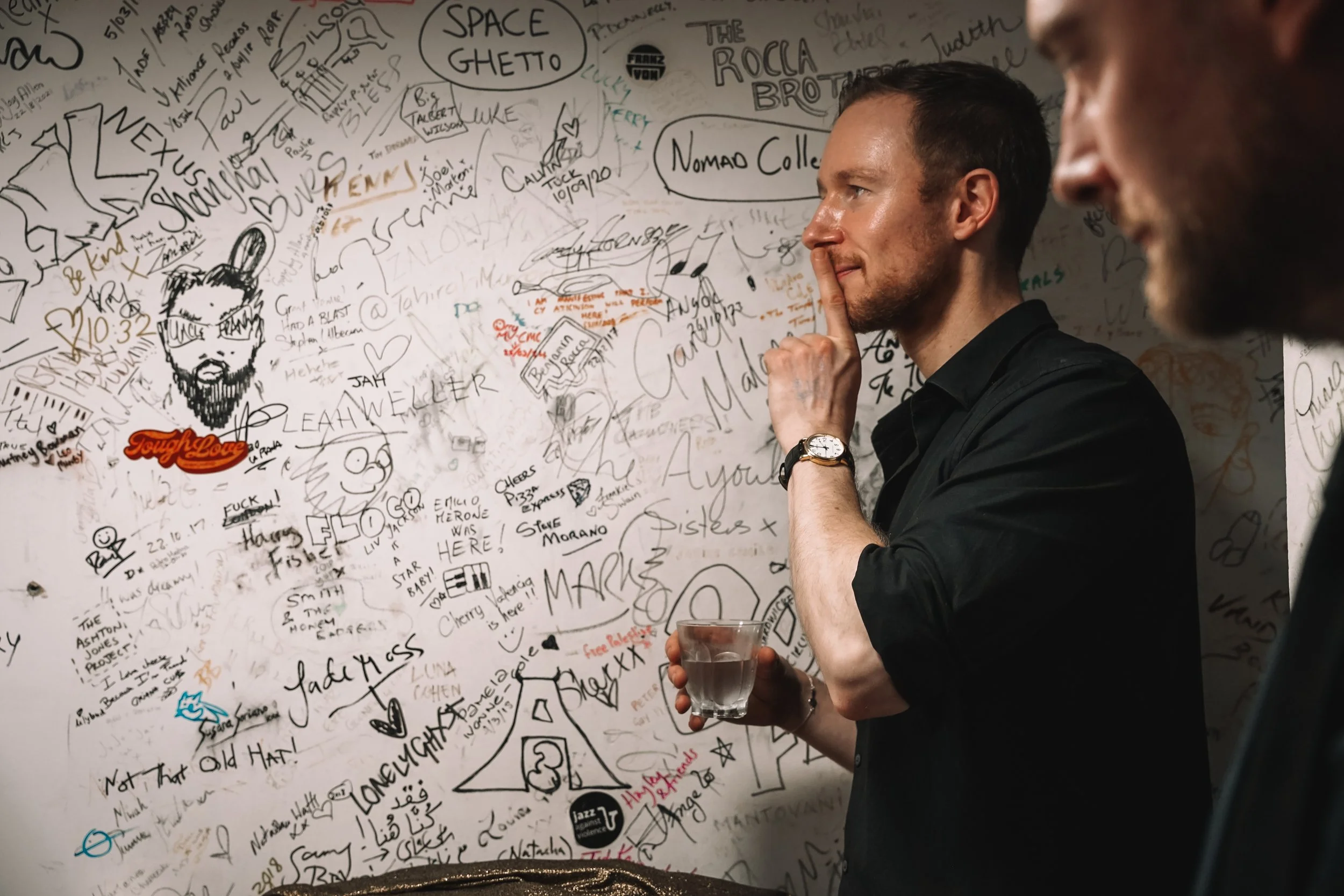 A man with short dark hair, wearing a black shirt and watch, is holding a glass and pressing his finger against his lips, standing in front of a wall covered in various signatures, drawings, and messages.