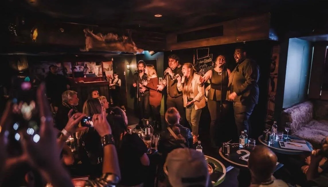Group of people singing on stage at a cozy bar or pub with an audience watching.