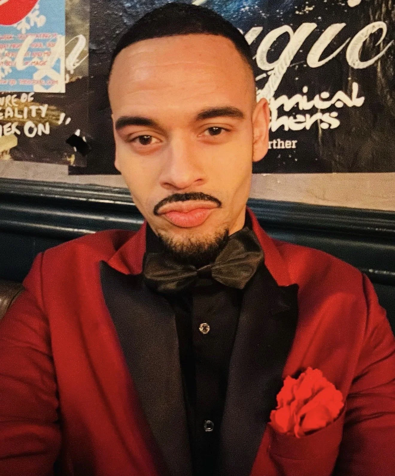 A man dressed in a red tuxedo with a black shirt, a black bow tie, and a red pocket square with a red flower. He has short black hair, a trimmed beard, and is making a duck face. Behind him is a blackboard with white writing and some colorful posters on the wall.